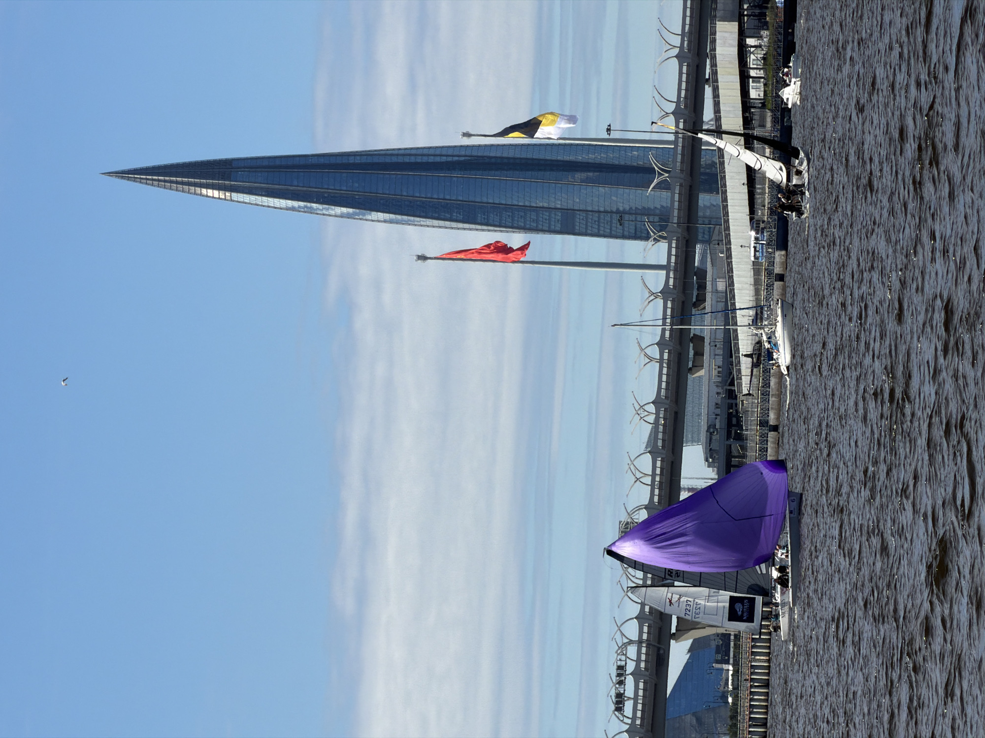 The image depicts a bright, sunny day with a clear blue sky, dotted with some wispy white clouds. In the foreground, a dark, rippled body of water is visible. Several sailboats are navigating the water. One prominent sailboat on the left has a large, vibrant purple sail, while another nearby has a white sail with black markings and the word "NAUTILUS" visible. Further to the right, another sailboat with a black and white sail is seen, with a few people on board. In the background, a distinctive, tall, pointed skyscraper with a glass facade dominates the skyline. This building is the Lakhta Center, located in Saint Petersburg, Russia. Two flags are flying from poles in front of the skyscraper: a bright red flag on the left and a black and yellow flag on the right. A large, elevated highway or bridge structure with distinctive arching supports crosses the scene horizontally behind the boats and in front of the buildings. The overall scene suggests a day of sailing near a modern urban waterfront in Saint Petersburg.