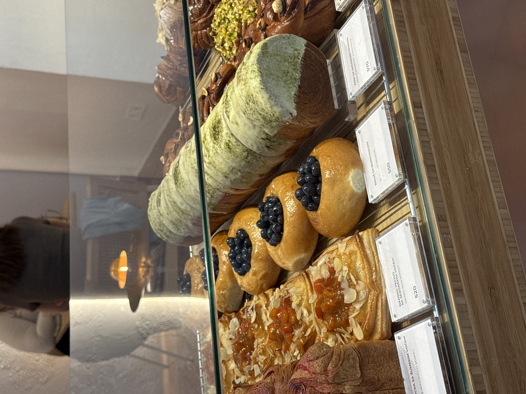 The image shows a display case filled with pastries in a bakery. In the foreground, several Danish pastries topped with apricot jam and almonds are visible. To the right of the Danish pastries are brioche buns filled with blueberries. A large, long pastry, possibly a roulade or a large croissant, with green matcha icing and white glaze is also prominent. In the background, croissants and other baked goods are displayed on shelves. A person's back is visible in the upper left portion of the image, suggesting they are a customer or employee. The display case has clear labels with text in what appears to be Serbian, including prices in the local currency. One label reads "DANSKO PECIVO SA SEZONSKIM VOCEM DANISH WITH APRICOT" with a price of "520". Another label says "BRIOS SA BOROVNICOM STRAWBERRY BRIOCHE" with a price of "550". A third label states "KOIN SA JAGODOM I MACOM CAKE STRAWBERRY AND MATCHA" with a price of "570". The lighting suggests an indoor setting, possibly a modern cafe or bakery. Given the Serbian text on the labels, the location is likely Belgrade, Serbia.