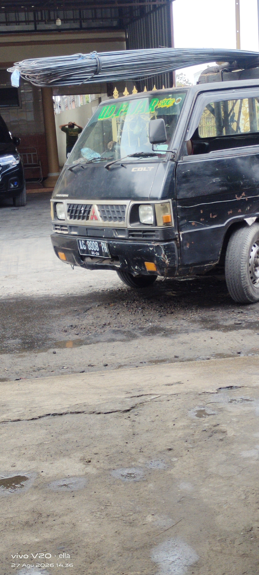 A black Mitsubishi Colt pickup truck is parked on a cracked asphalt surface, likely in Simo, Indonesia, under a covered structure. The truck is loaded with a large bundle of long, thin metal rods strapped together with blue twine, which extends over the cab and is secured to the vehicle's roof rack. The truck's front grille displays the Mitsubishi logo. The license plate reads "AG 8908 PH". A person wearing a green shirt and dark pants is visible in the background, facing away from the camera and appearing to take a photo. The area around the truck is wet with puddles, suggesting recent rain or damp conditions. A portion of a black SUV is visible to the left of the truck. The overall scene depicts a commercial or logistical operation involving the transport of construction materials.