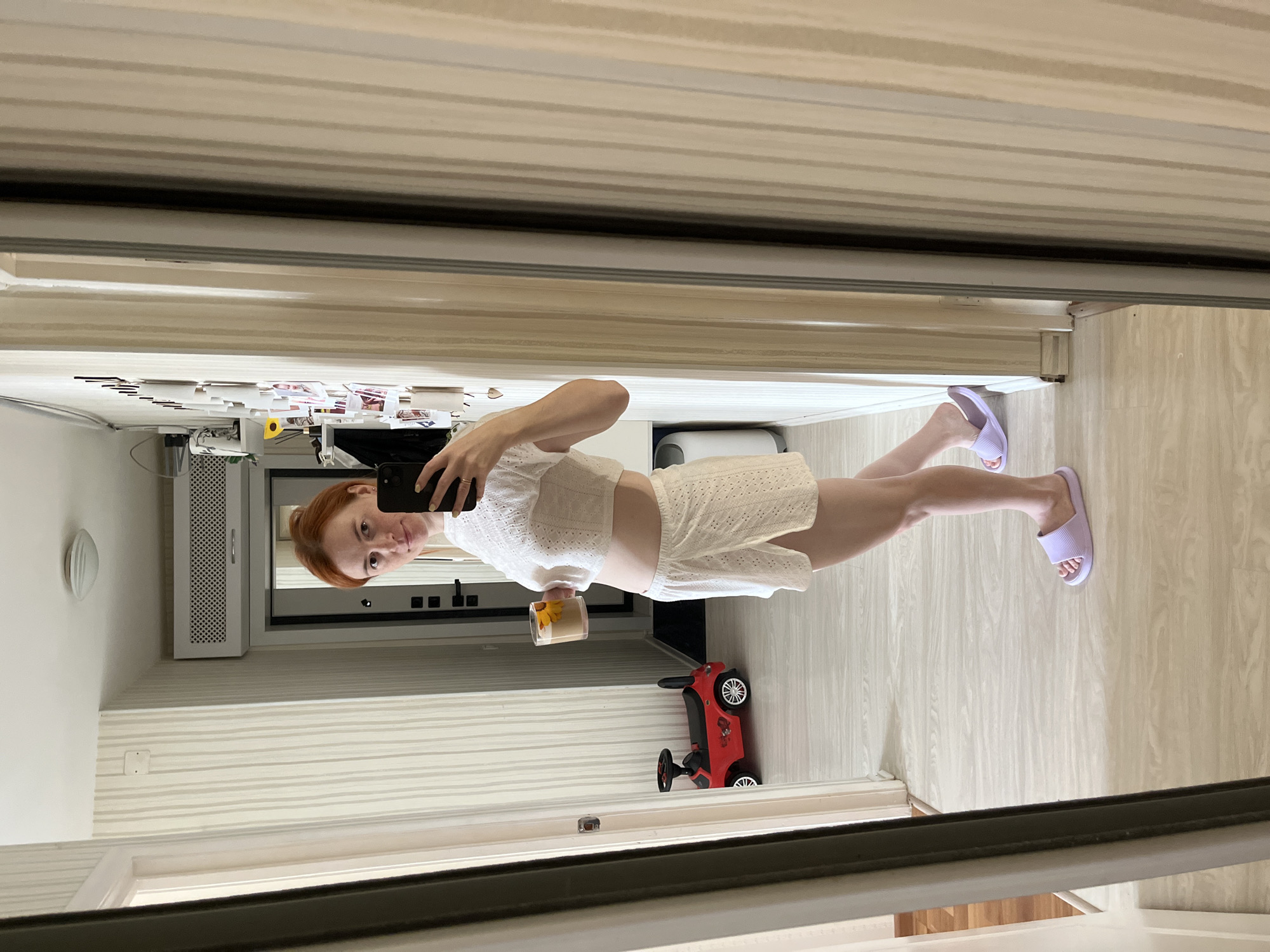 A woman with red hair is standing in a hallway, taking a mirror selfie. She is wearing a white crop top and white shorts, and purple slides. She is holding a cup of coffee. In the background, there is a red toy car, a door, and a wall with vertical striped wallpaper. The image appears to have been taken in an apartment in Chaikovsky, Russia.