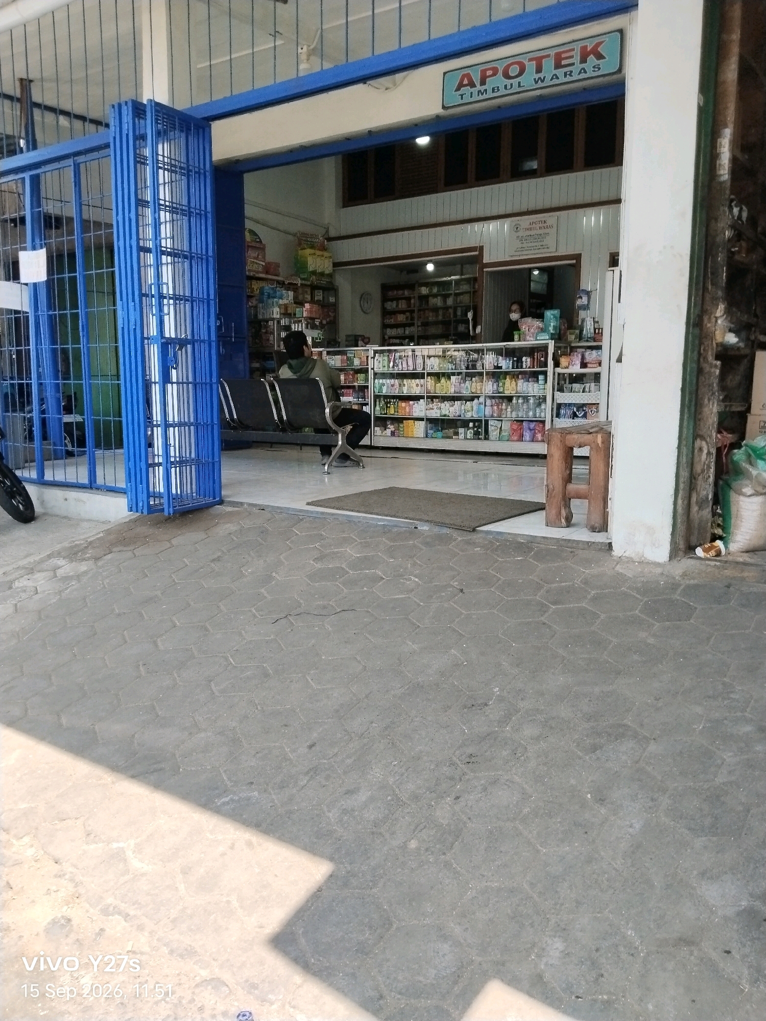The image shows the exterior of a pharmacy named "APOTEK TIMBUL WARAS" located in Sukorejo, Indonesia. The pharmacy has a blue-framed entrance with folding doors. Inside, shelves are stocked with various products. A person is seated on a black bench near the entrance. Another person wearing a mask is visible behind a counter inside the pharmacy. The ground in front of the pharmacy is paved with hexagonal concrete tiles. Sunlight casts shadows on the pavement. The timestamp on the image indicates it was taken on September 15, 2026, at 11:51 AM.