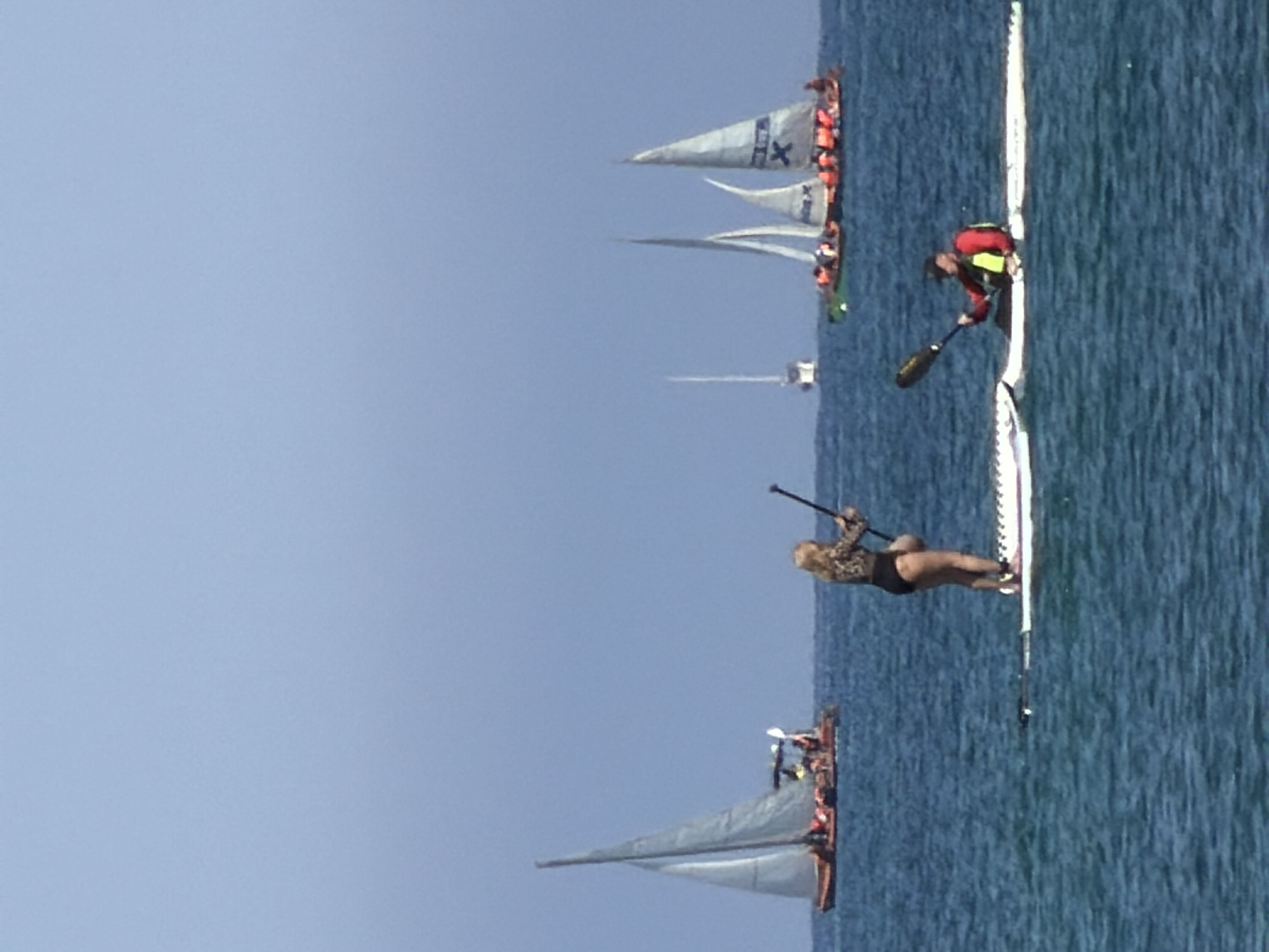 The image shows a water scene in Tel-Aviv, Israel. There are several dragon boats with sails and multiple people aboard, wearing orange life vests. Two individuals are on a long, narrow paddleboard; one is kneeling and paddling, while the other is standing and also paddling. The water is blue, and the sky is clear.