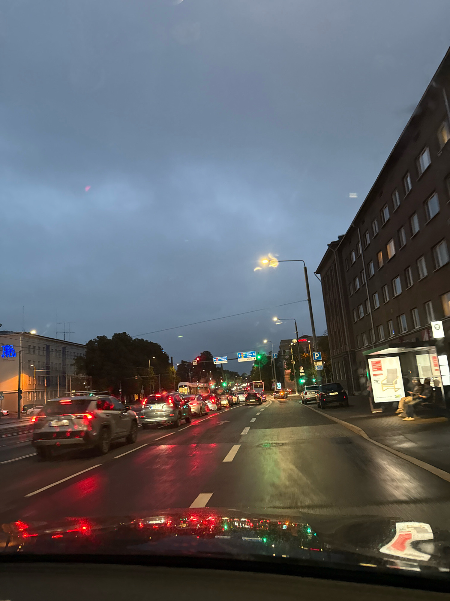 The image depicts a street scene in Tallinn, Estonia, at dusk, with heavy traffic. The perspective is from inside a vehicle, looking forward. The road is wet, reflecting the red taillights of cars and green traffic signals, suggesting recent rain or damp conditions.
On the right side of the road, a multi-story building with numerous windows is visible. A bus stop shelter with an advertisement featuring a chair and a price tag of "79€" is also present. Two individuals are seated at the bus stop.
On the left side of the road, another building with illuminated windows is seen, along with a sign displaying blue neon lettering that reads "TEC TINO." Further down the street, traffic lights are visible, showing red and green signals. Multiple cars are lined up, indicating congestion. Trees line the street in the middle distance. The sky is overcast and dark, characteristic of evening or a cloudy day. A street lamp with a yellow light fixture illuminates the scene.