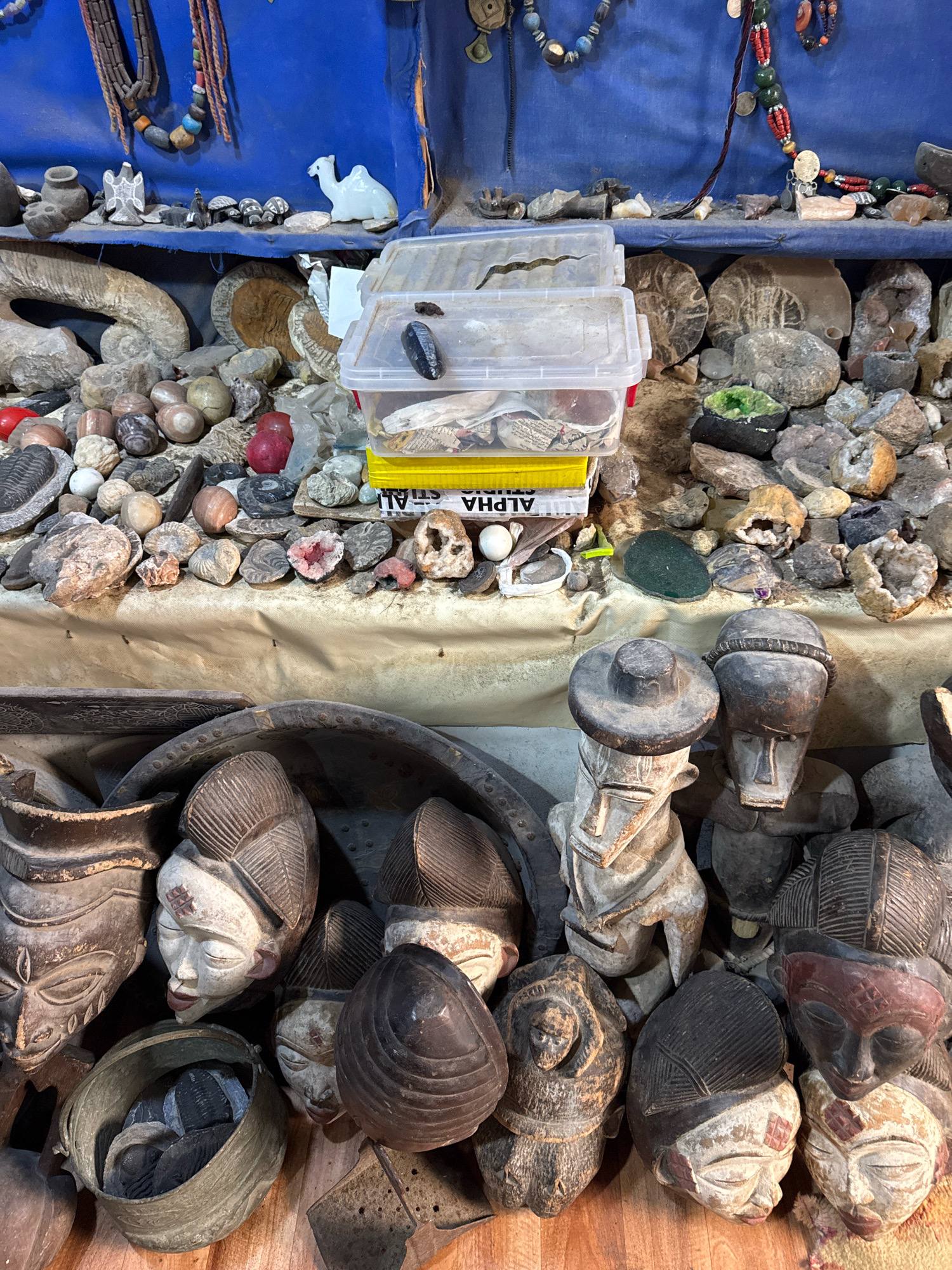 The image depicts an indoor stall likely located in Essaouira, Morocco, exhibiting a variety of artisanal and natural items. The scene is densely packed with objects displayed on shelves and on a lower surface.
The upper shelf is covered with a blue fabric backdrop and displays various necklaces, beads, and carved items. Notable objects include what appear to be fossilized shells, small animal figurines including a white camel, and various stones and rocks. Two clear plastic containers are also present, one stacked on top of a yellow box, containing what looks like papers and possibly more stones.
The lower surface is a densely arranged collection of items. In the foreground, there are several carved wooden masks and figures. These appear to be of African origin, with intricate details and varied expressions. Some masks are painted or have remnants of paint. Interspersed among the masks are various stones, fossils, and what appear to be geodes or crystalline rock formations. One prominent large, dark bowl-like object contains several smaller, dark, possibly fossilized items.
The lighting suggests an indoor setting, possibly a market stall or a private collection. The overall impression is one of a curated display of natural curiosities and handcrafted artifacts.