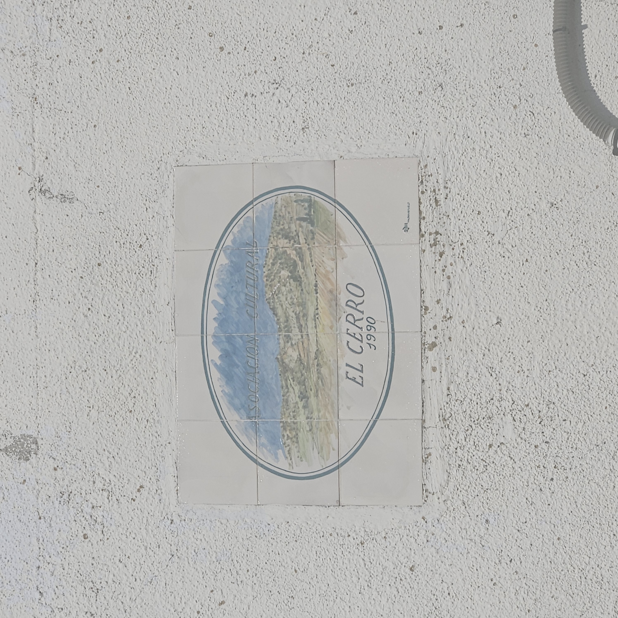 A tiled plaque is mounted on a white, rough textured wall. The plaque consists of 6 tiles, arranged in a 2x3 grid, depicting an oval scene. The scene shows a mountain landscape with blue water or a lake to the left, and a mountainous region in the center and right. The words "ASOCIACION CULTURAL" curve along the top left of the oval. The words "EL CERRO" and "1990" are on the right side of the oval. The plaque appears to be a commemorative or promotional sign related to the town of El Cerro, perhaps a cultural association established in 1990. The plaque's style is reminiscent of traditional Spanish ceramic tiles. There is a utility conduit on the top right side of the wall. Based on the title, this is in Algora, Spain.