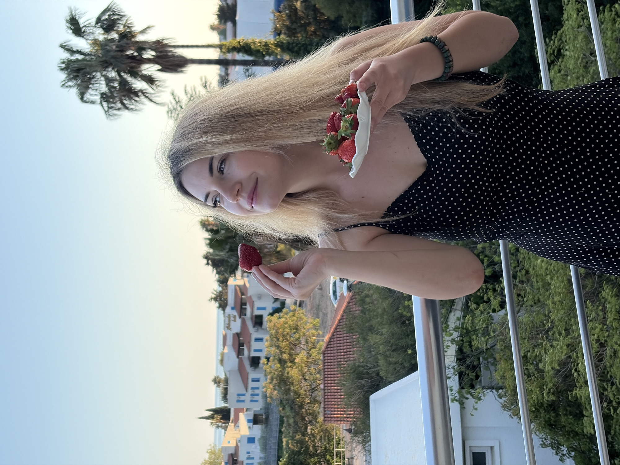 A woman with long blonde hair holds a small plate of strawberries in one hand and a single strawberry in the other. She is wearing a black polka-dot dress and a bracelet. She is standing on a balcony with metal railings. Behind her, there are white buildings with blue windows and red tile roofs. Green trees and a palm tree are visible. The location appears to be Budva, Montenegro, based on the architectural style of the buildings. The sky is a pale blue color, suggesting it is either early morning or late afternoon.