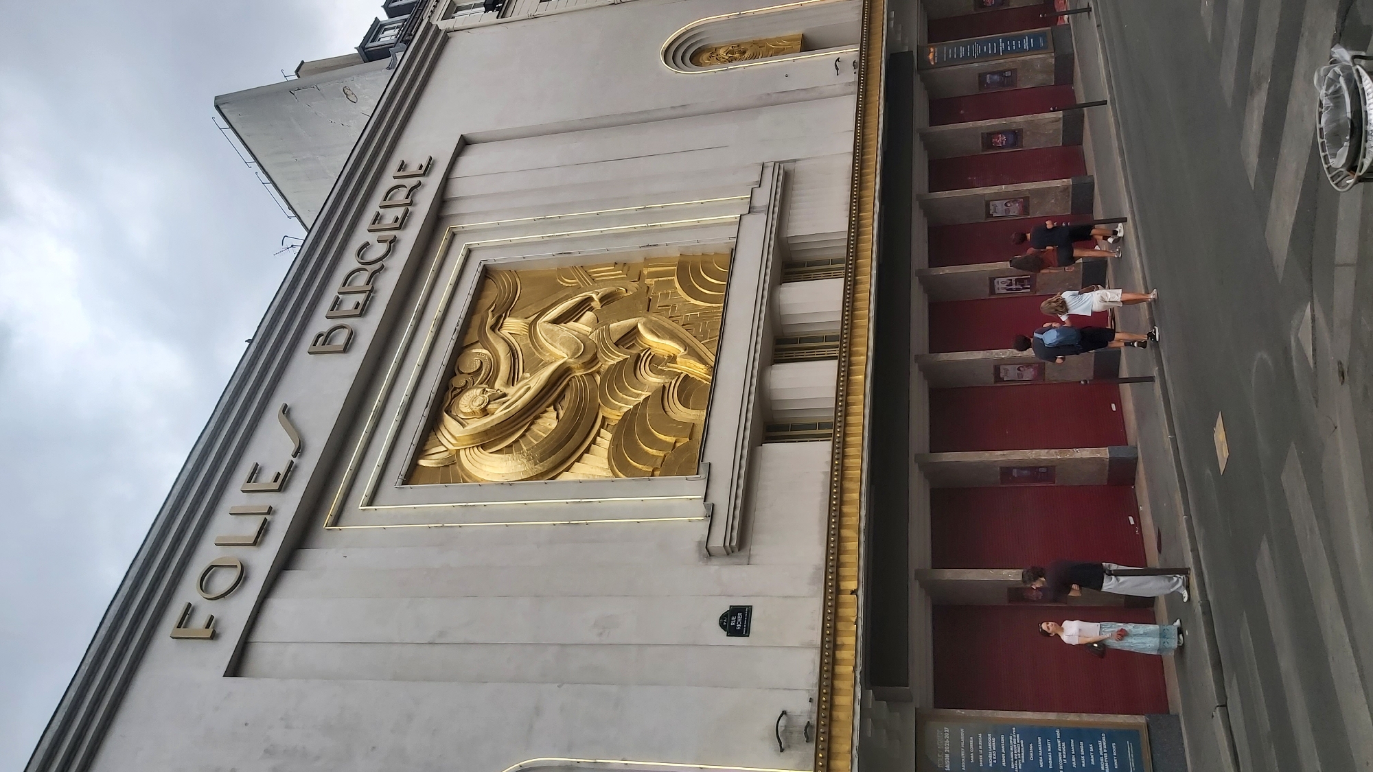 The image shows the Folies Bergère theater in Paris, France. The facade of the theater is light gray with gold lettering spelling out "FOLIES BERGERE" above a large, ornate gold relief sculpture of a dancer. Below the sculpture are several large, dark red doors. A street runs in front of the theater, with a crosswalk in the foreground. Several people are walking on the sidewalk in front of the theater, some looking at a poster board. The sky is overcast.