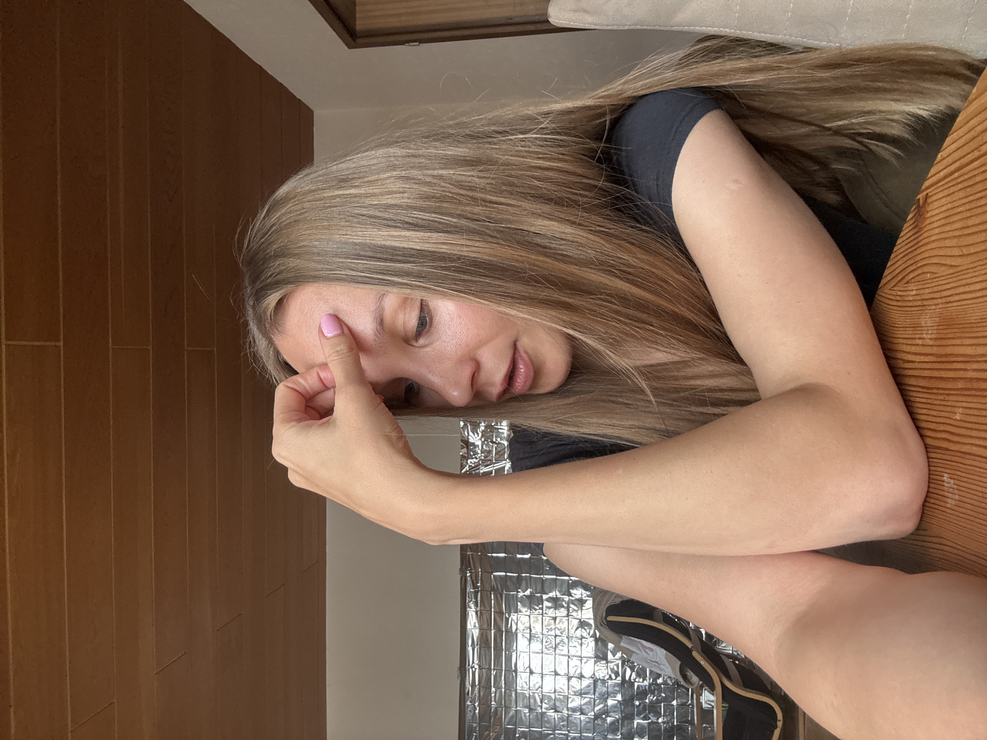 A young woman with long blonde hair is sitting at a wooden table, resting her forehead with her right hand. She is wearing a dark t-shirt. The background shows a wooden ceiling and a wall with a silver, reflective surface. A chair is visible to the right of the woman. The image appears to be taken indoors, possibly in a home or a restaurant. There is no discernible connection to Überherrn, Germany.