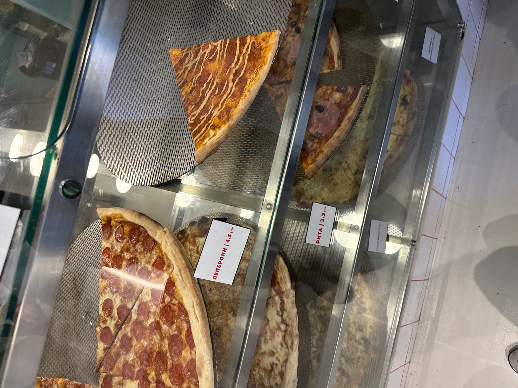 The image shows a display case filled with pizza slices, likely in a restaurant or fast food establishment in Sofia, Bulgaria. There are several different varieties of pizza visible, including pepperoni, a pizza with ham and olives, and a pizza topped with a sauce resembling a glaze. Each pizza slice has a label indicating its type and price in euros. The display case is well-lit, and the pizzas appear fresh. The overall impression is of a quick and convenient food option in an urban setting.