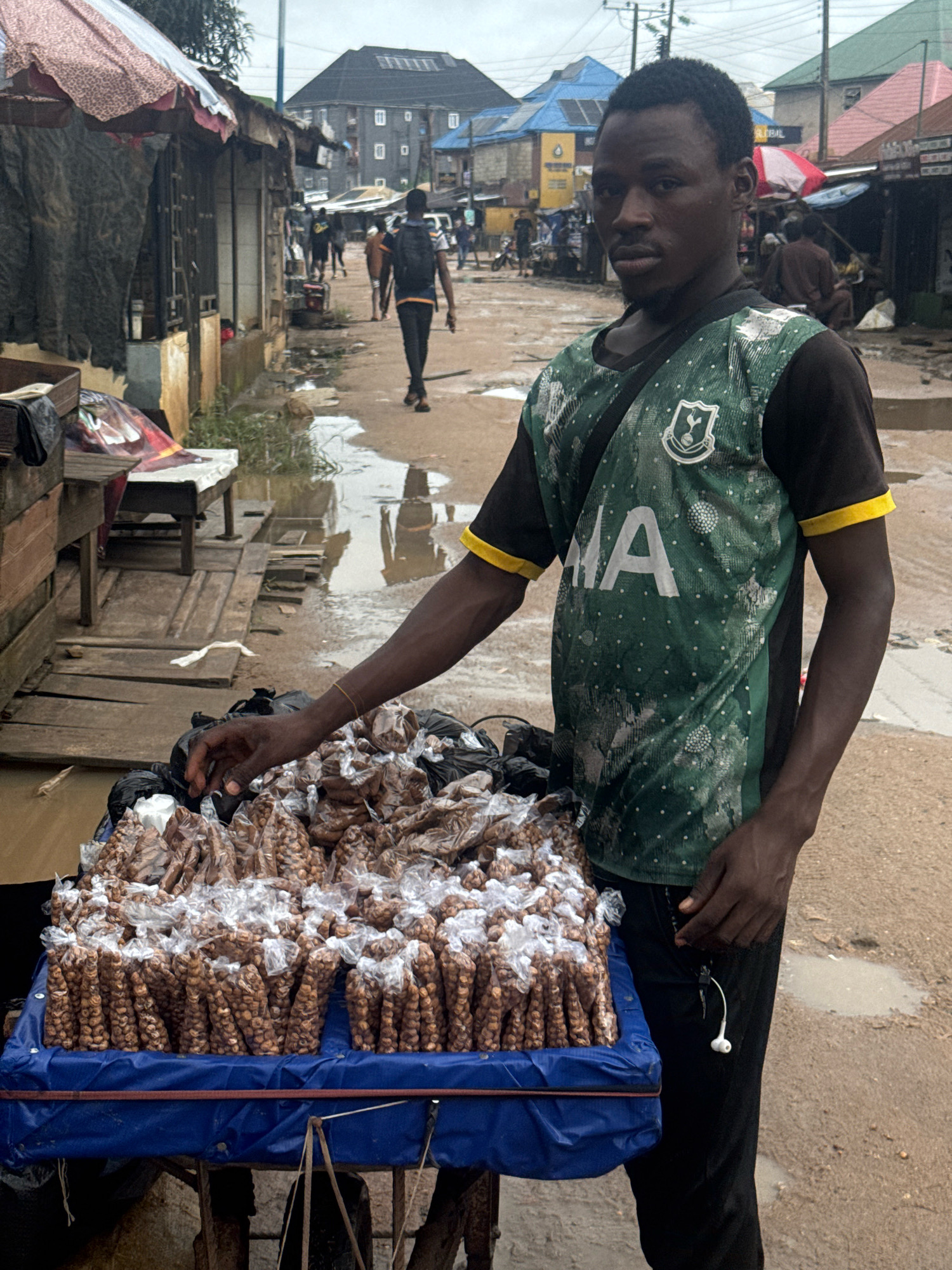 A young man stands behind a display of packaged nuts on a cart on a muddy street in Ihiagwa, Nigeria. He is wearing a green and black soccer jersey and black pants. His left hand is reaching towards the nuts. The street is wet and appears to have puddles of water. In the background, there are several market stalls and buildings. Other people are walking in the distance. The sky is overcast.