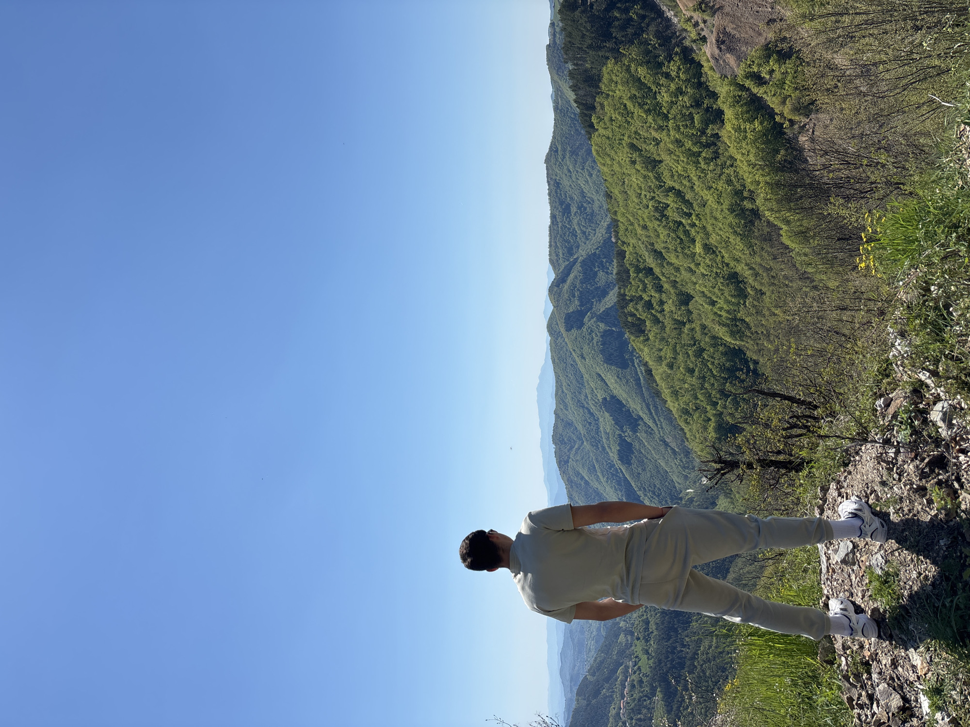 The image shows a man standing on a rocky cliff overlooking a mountainous landscape near Petrov Dol, Bulgaria. The man is wearing light-colored clothing and white sneakers with blue accents. He is facing the valley, looking out at the forested hills and the blue sky. The foreground shows the edge of the cliff with some vegetation. The scene is captured on a bright, sunny day.