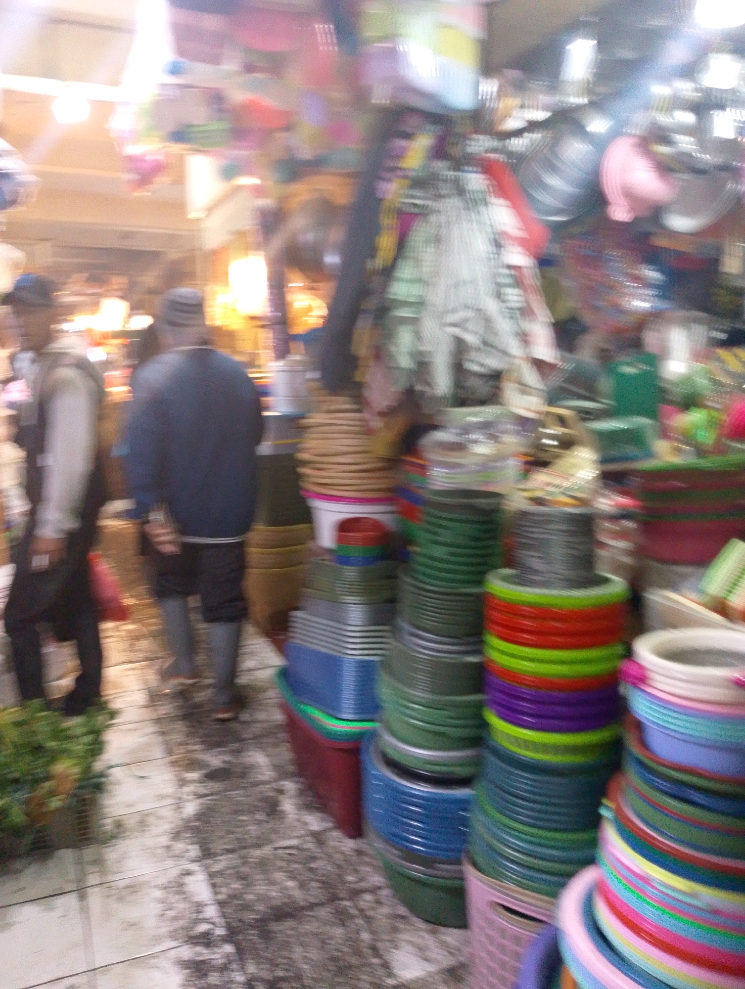 The image depicts a bustling market scene in Lembang, Indonesia, likely an outdoor or semi-outdoor setting given the lighting and open feel. The foreground is dominated by stacks of colorful plastic containers and baskets in various sizes and hues, including green, blue, red, purple, and yellow. These are arranged in a somewhat organized but dense manner, suggesting a vendor's stall.
In the mid-ground, two individuals are visible. On the left, a person wearing a light-colored jacket and dark pants walks away from the camera, their face obscured. To their right, another person stands with their back to the viewer, wearing a dark blue long-sleeved shirt and dark pants, with a striped cap on their head. Their posture suggests they might be a vendor or a shopper.
The background is blurry, but appears to show more stalls with merchandise, possibly including textiles or household goods. The lighting is bright and somewhat diffused, creating a slightly overexposed effect in some areas, especially towards the top of the image. The ground surface seems to be a mix of tiled and possibly damp or worn concrete. The overall impression is one of lively commerce and activity within a typical Indonesian market environment.