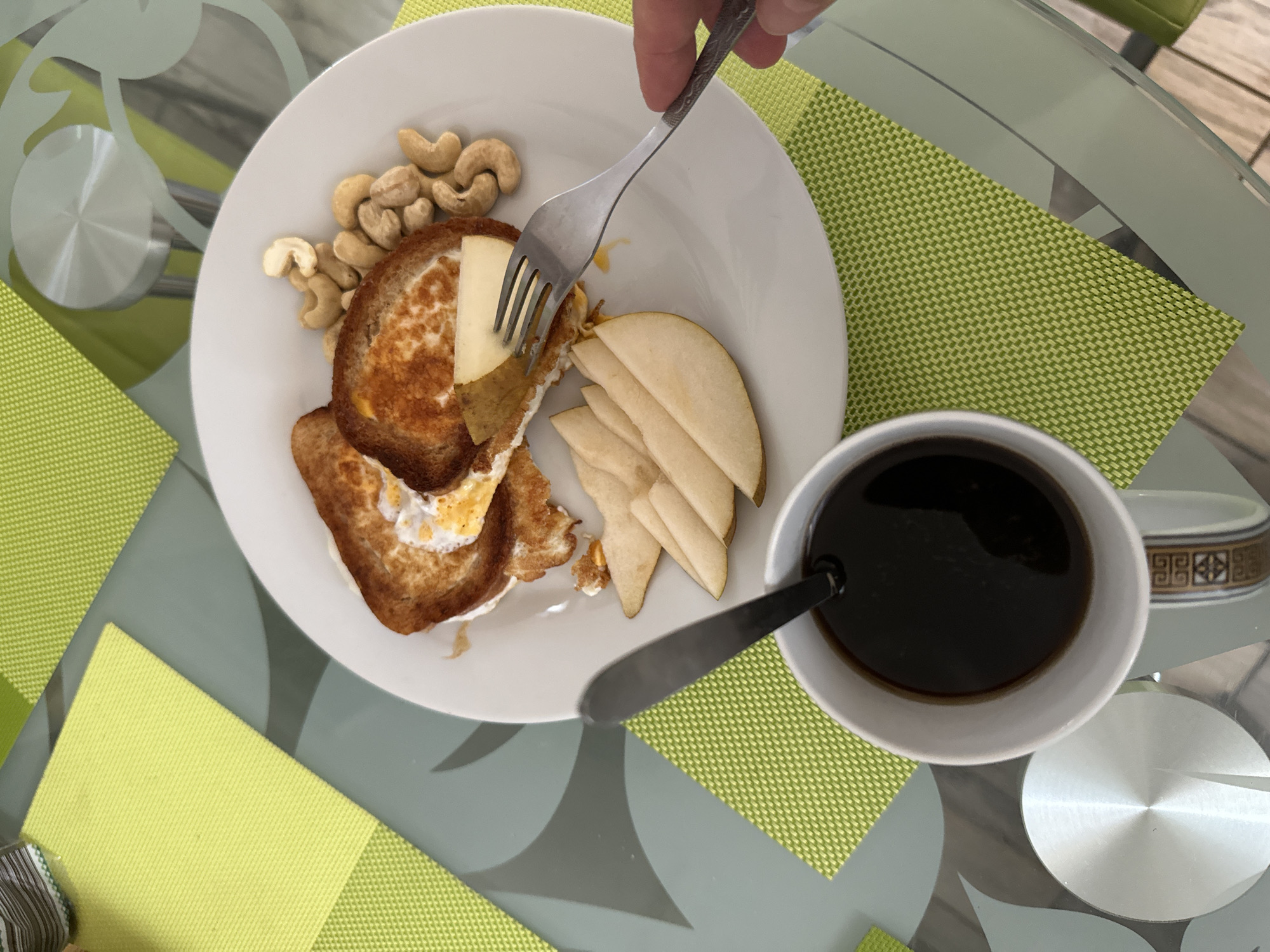 The image depicts a breakfast scene on a glass table, set with a white plate containing toasted bread with egg and cheese, sliced pears, and cashew nuts. A white mug filled with dark liquid, presumably coffee, is placed to the left of the plate, with a spoon inside. A fork, held by a person's hand which is partially visible on the right side of the frame, is in the process of picking up a slice of pear from the plate. The table is adorned with lime green placemats. The background shows a reflective glass surface with a subtle pattern. No specific location in Podolsk, Russia, can be identified from the visual cues in the image.