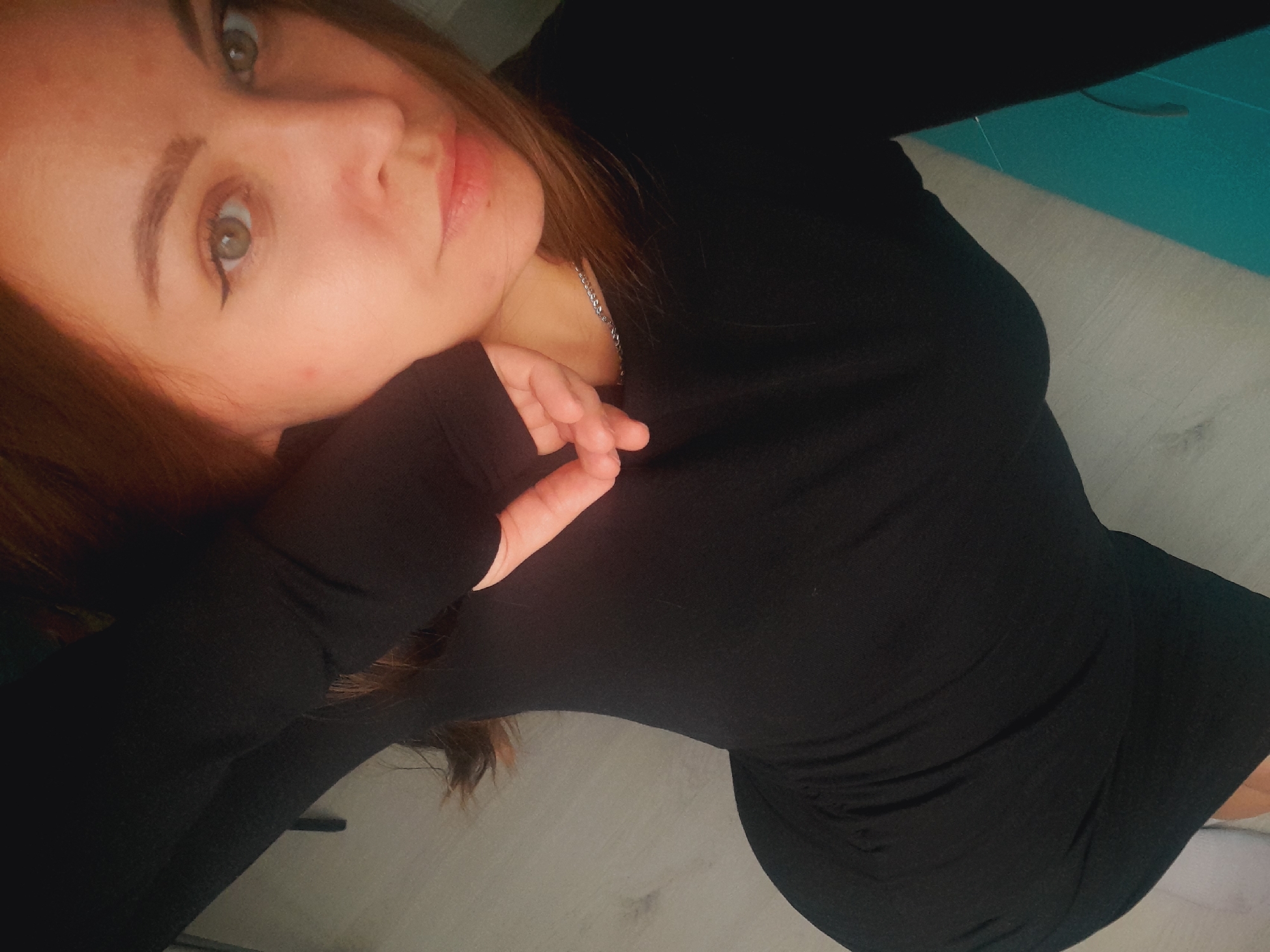 The image shows a woman with light skin, light brown hair, and green eyes, angled slightly to the left. She is wearing a black long-sleeved dress or top. Her left hand is positioned near her face. A thin silver necklace is visible on her neck. The background includes a teal or turquoise-colored object near the top right, a light gray surface that looks like a floor, and a small glimpse of what appears to be a white object or surface near the bottom right corner. The location in the city of Unknown, Unknown is not discernible from the image.
