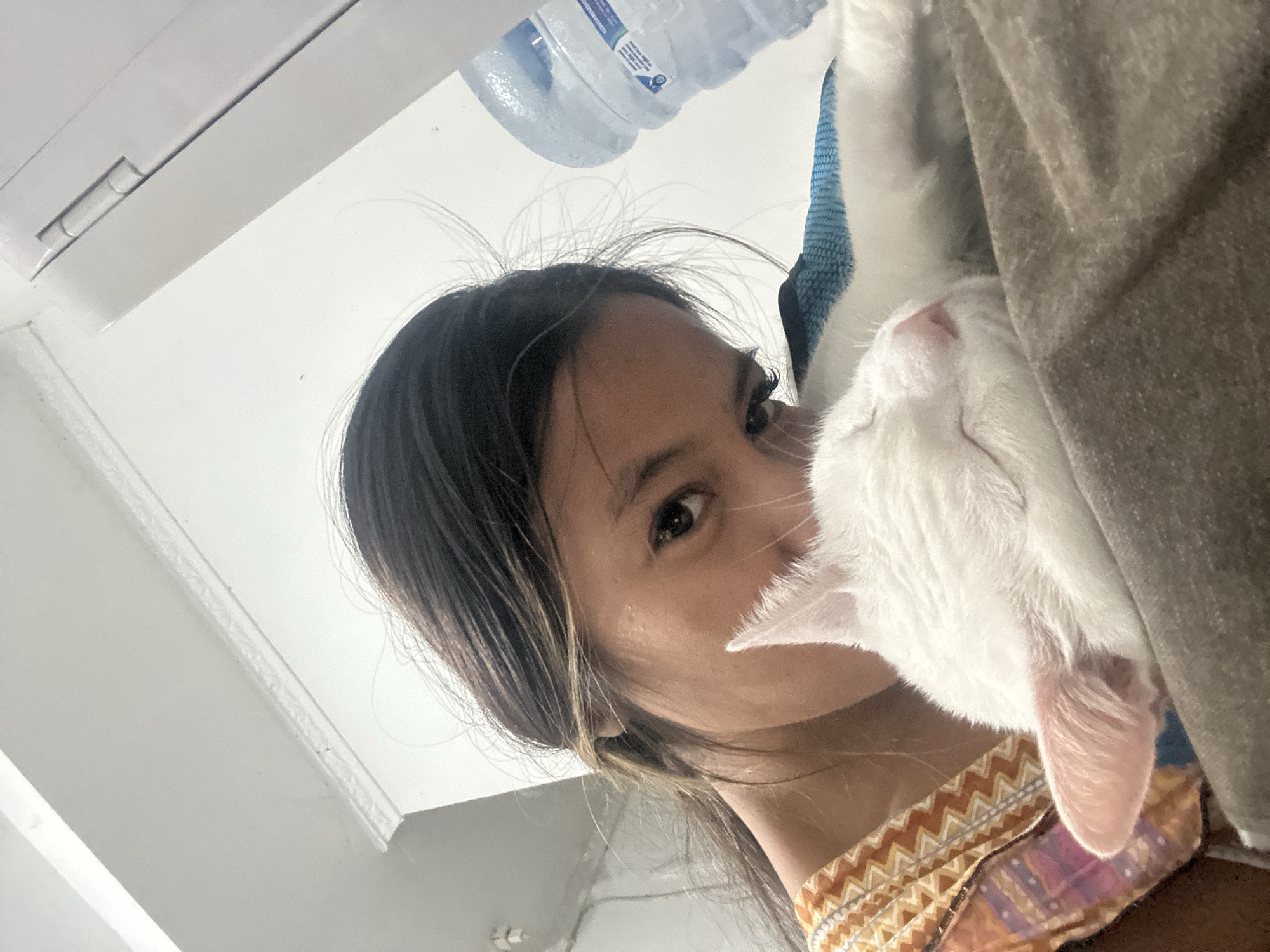 A woman with dark hair and brown eyes is holding a white cat to her face. The cat's head is against the woman's cheek. A plastic water bottle is visible above the woman's head. The background includes a white ceiling and a gray fabric. The woman is wearing a patterned shirt. The location is potentially North Jakarta, Indonesia.