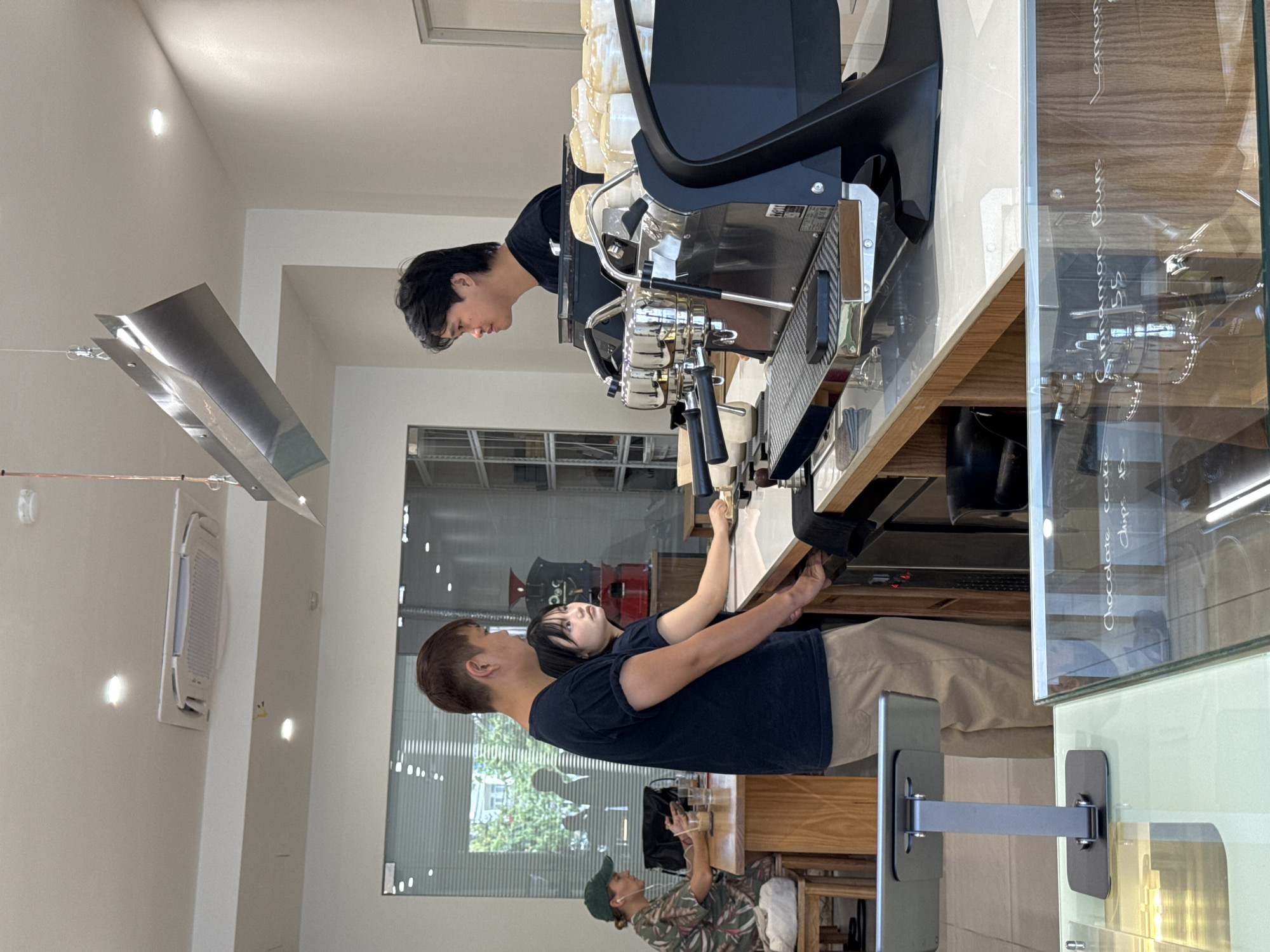 The image depicts the interior of a modern cafe in Da Nang, Vietnam. Three individuals are visible. In the foreground, a man in a dark blue t-shirt and beige pants stands behind a counter, facing a young woman with short dark hair. The woman is wearing a dark t-shirt and is positioned to his right, appearing to interact with him. To the left, a woman wearing a green cap and a patterned shirt sits at a wooden table, looking at her phone and wearing earbuds. Behind her, a coffee machine with chrome accents and a black casing is prominent on the counter. The cafe's interior features a clean, minimalist design with white walls and wooden accents. A large window reflects the outside environment. In the foreground, a glass display case shows baked goods with handwritten labels such as "Chocolate cookie" and "Cinnamon buns." The overall scene suggests a casual atmosphere within a food and beverage establishment.