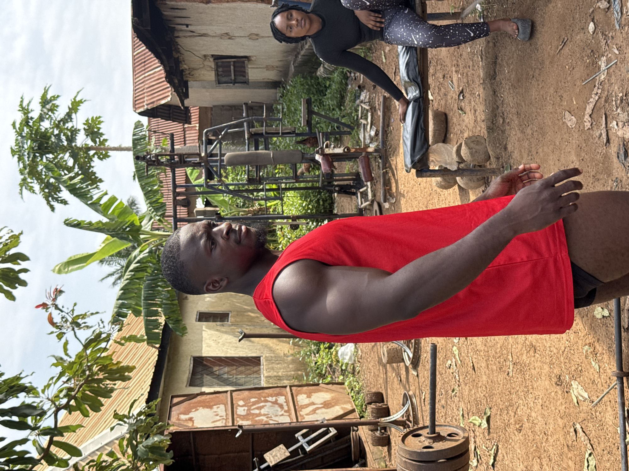 A medium shot captures a muscular Black man in a red tank top and dark shorts, facing right and looking off-camera. He stands in what appears to be an outdoor gym in Akwanga, Nigeria, with a woman in a black long-sleeved shirt and patterned leggings seated on a bench behind him to the right. The gym equipment, including a weight rack with rusted plates and a bench press, is visible. The background features rustic buildings with corrugated iron roofs, overgrown vegetation, and dirt ground, suggesting a rural or semi-urban setting. The lighting indicates it is daytime with an overcast sky.