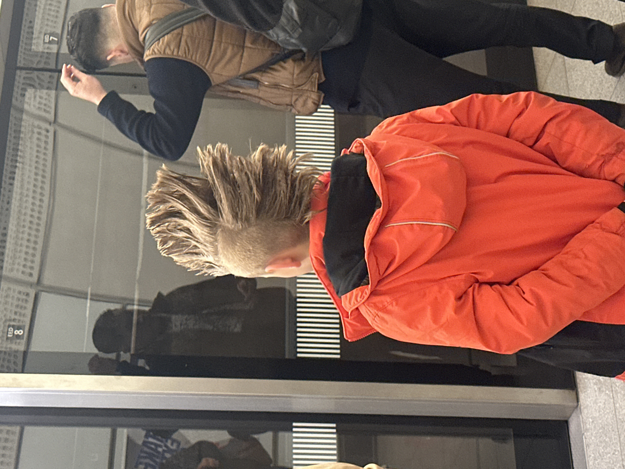 Two individuals are visible in the frame. One, wearing an orange jacket, faces towards the right. The second, clad in a brown vest over a dark long-sleeved shirt and dark pants, faces towards the left. The subjects are reflected in a window displaying the number "8". This suggests they are inside an elevator. The precise location cannot be determined from the provided image.
