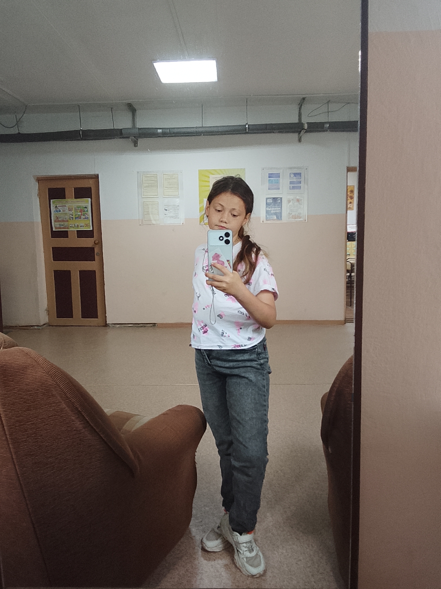 In the city of Chaikovsky, Russia, a young girl with brown hair pulled back into a ponytail stands in front of a large mirror, taking a selfie with her phone. She is wearing a white t-shirt with a pink floral and flamingo pattern and dark grey jeans. She has on white sneakers with orange accents. The girl is positioned in what appears to be an indoor hallway or common area, possibly a school or community center, given the bulletin boards with papers on the wall behind her. A closed wooden door with decorative panels is visible to her left, and a poster with colorful illustrations is affixed to it. To the right of the girl, a large mirror dominates the frame, reflecting the scene and the girl herself. The floor is a neutral tiled surface. Two large, brown, upholstered chairs are partially visible in the foreground, suggesting a seating area. The lighting appears to be from overhead fluorescent fixtures.