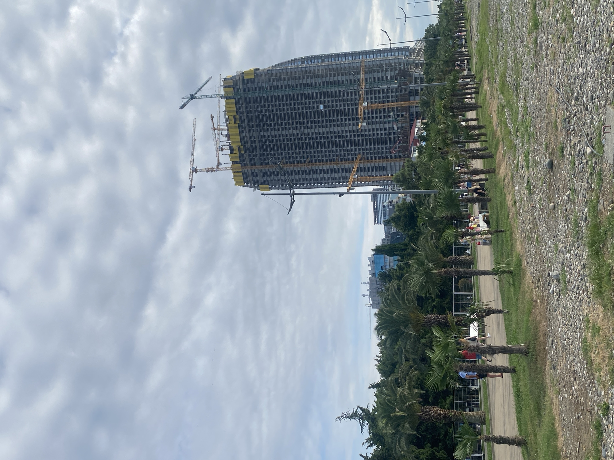 The image captures a construction site in Batumi, Georgia, featuring a partially completed skyscraper. The skyscraper, a tall, multi-story building with a dark facade, is surrounded by construction cranes. The area surrounding the building is lined with palm trees, suggesting a coastal or tropical environment. Several people are visible walking along a paved path next to the palm trees. The ground in the foreground consists of gravel and stones. The sky is overcast with clouds.