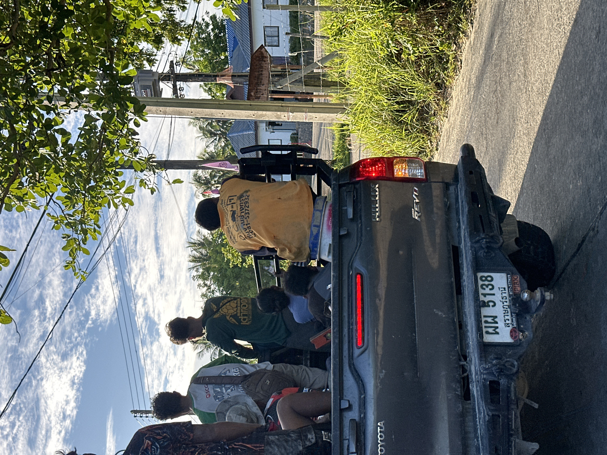 The image shows the rear of a dark gray Toyota Hilux Revo pickup truck, with several people in the bed. The license plate reads "พพ 5138" and "สุราษฎร์ธานี". The truck appears to be on a paved road with a grassy embankment to the right. In the background, there are houses with blue roofs, palm trees, and electrical poles with wires. The sky is blue with scattered white clouds, and a tree with green leaves is visible in the upper right corner. The people in the truck are facing away from the camera, and their clothing suggests they might be workers. One person in the center is wearing an orange shirt with text on the back. The overall scene suggests a rural or semi-rural setting in Thailand, consistent with the location "ตำบลเกาะพะงัน, Thailand".