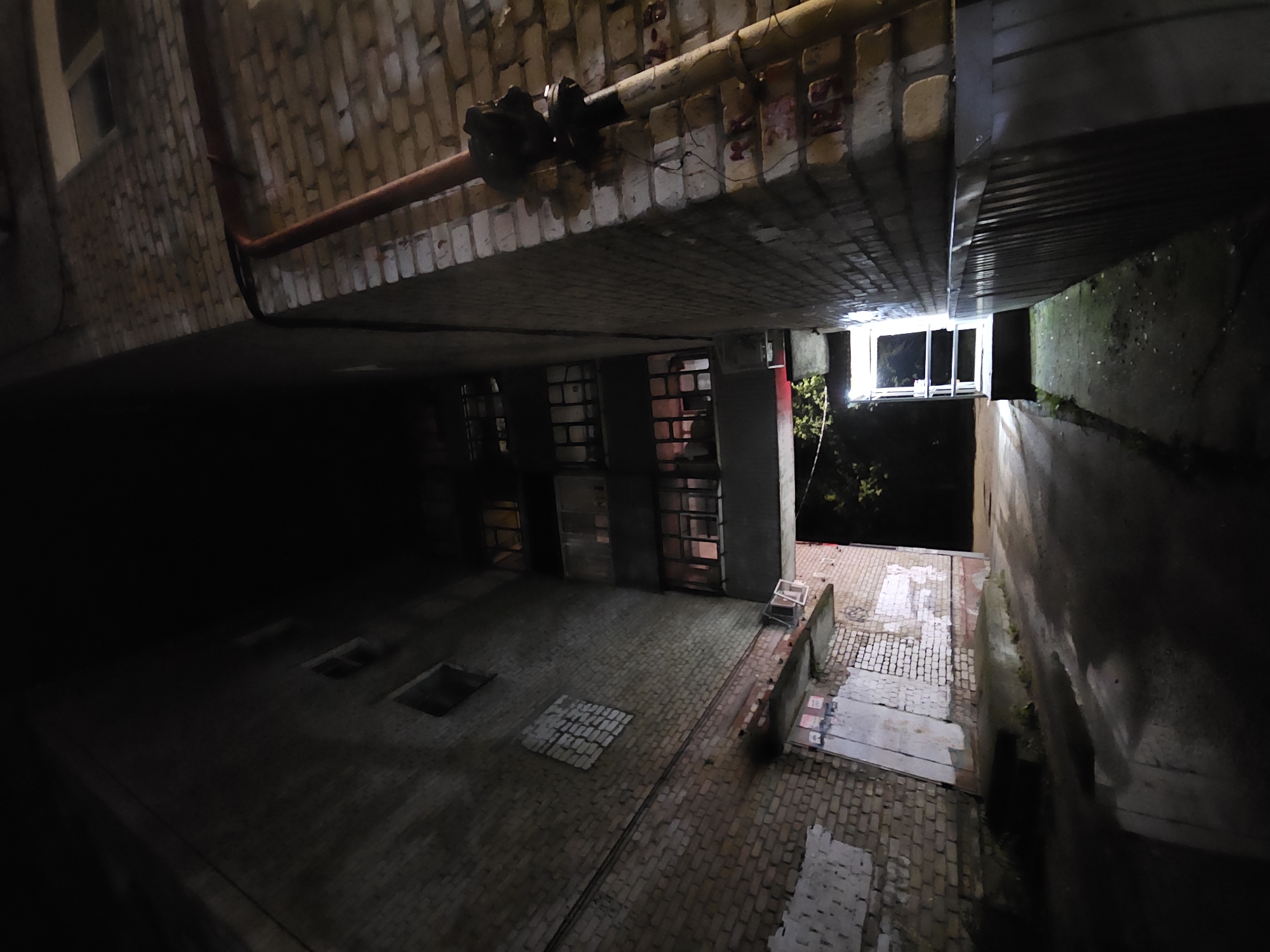 This image depicts a dimly lit alleyway between two brick buildings at night. The perspective is looking upwards, emphasizing the height and narrowness of the space. The building on the left is a multi-story brick structure with several windows, some appearing to be dark, and others with a faint glow. A section of the brickwork on this building shows a patch of white material. The building on the right is also brick and features a prominent red pipe running vertically along its exterior, connected to a larger apparatus. This building has a gray, ribbed surface at its base.
The alleyway itself is paved with asphalt and appears wet, reflecting the limited light. In the background, at the end of the alley, is an entrance with bright interior lighting and a metal railing. Trees are visible to the left of this entrance. The overall atmosphere is dark and somewhat utilitarian. The location is identified as Ulyanovsk, Russia.