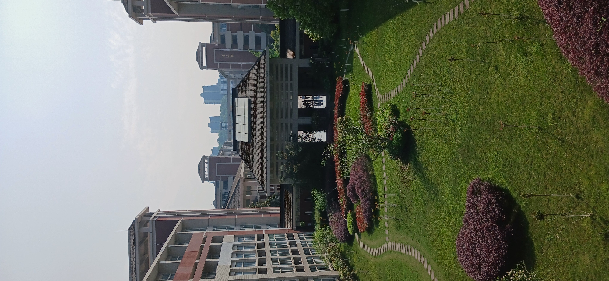 The image depicts a view of Chongqing High-tech Zone, China. In the foreground, there is a green lawn with winding stone paths. Various bushes and trees are planted along the paths. Buildings of varying heights are visible in the background. The sky is overcast with a slight haze.