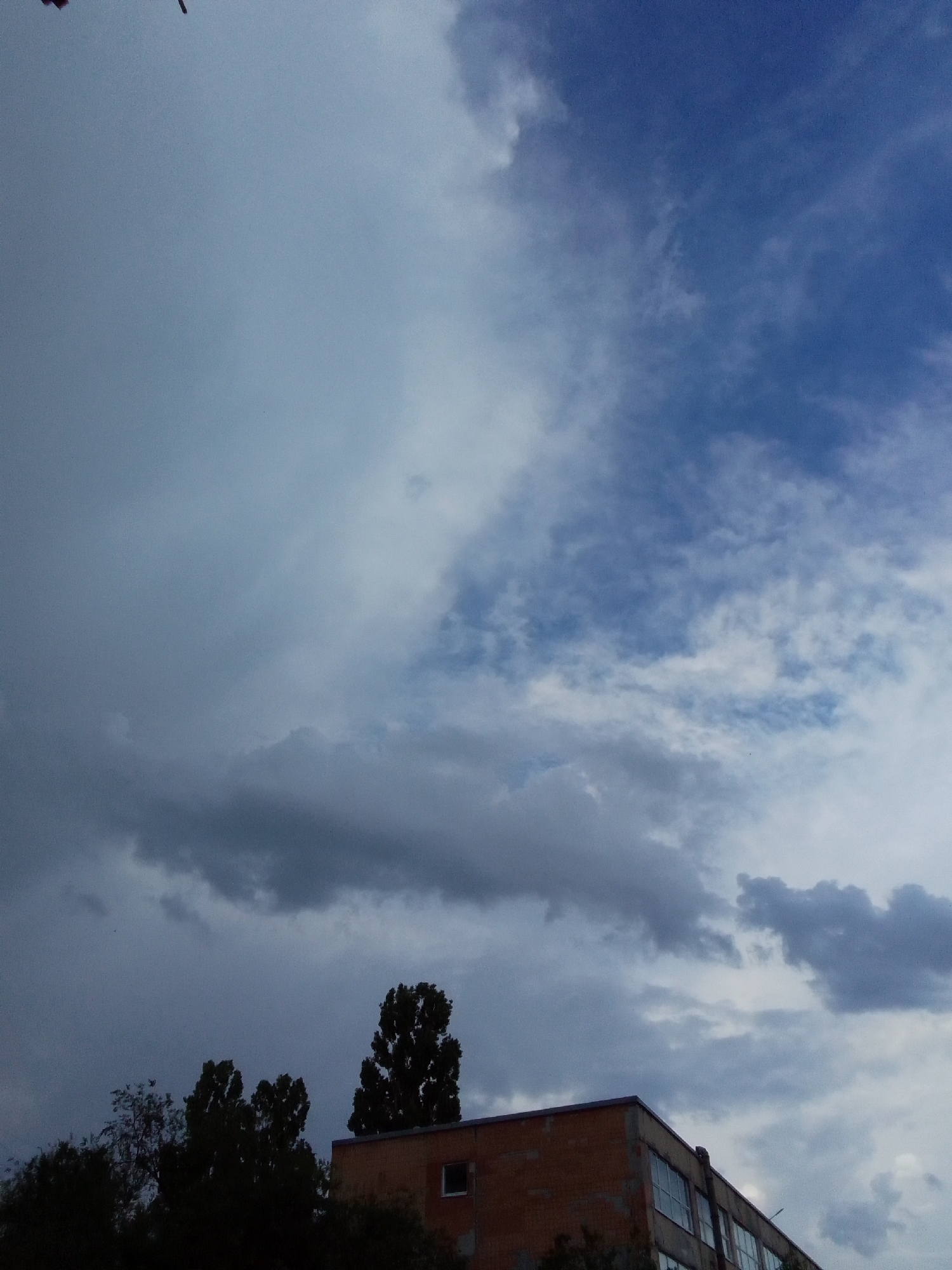The image captures an outdoor scene in a city. The sky is predominantly filled with clouds, ranging from dark, stormy-looking formations on the left to lighter, more scattered clouds on the right, revealing patches of blue sky. In the lower portion of the frame, a building is partially visible. It appears to be constructed of brick and has several windows. To the left of the building, several trees are silhouetted against the sky, their dark foliage contrasting with the lighter sky. The overall impression is that of an overcast or approaching storm day.