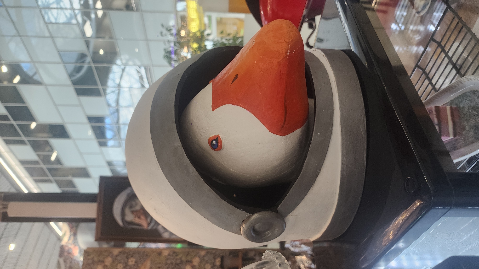 A goose wearing a white and gray astronaut helmet is positioned on a black reflective surface. The goose's head is visible within the helmet's opening, with its orange beak protruding. Its eye is depicted as a small blue circle with a red outline. The background shows a modern building with a grid-like facade of windows, reflecting the sky and interior lights. In the distance, partially obscured, is a framed picture of a person in an astronaut helmet. The scene appears to be indoors, possibly in a public space or a retail environment. The location is not specified in the image.