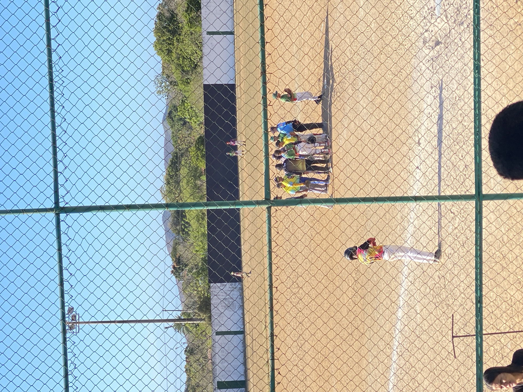 A group of people are gathered on a baseball field in Juan Griego, Venezuela, as seen through a chain-link fence. Some players are wearing green, yellow, and blue jerseys, while one player is wearing a brightly colored jersey. One individual stands apart from the group, and another walks nearby with a glove. The environment consists of a dirt infield, a green and white outfield fence, and trees visible behind the outfield fence. The sky is clear and blue. The time of day appears to be daytime due to the presence of shadows.