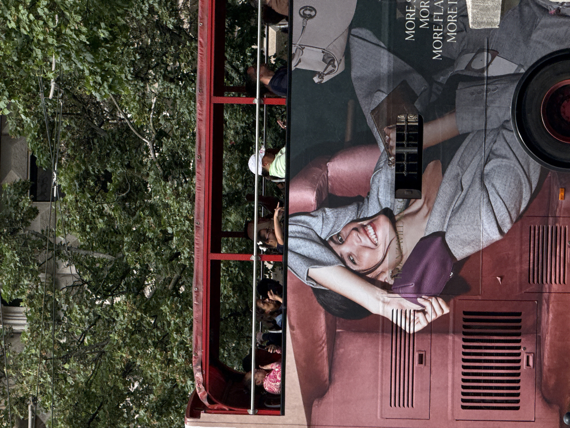 This media file captures a side view of a red double-decker bus, likely a tour bus, positioned against a backdrop of lush green trees and a building. The image is rotated 90 degrees counter-clockwise from its upright orientation.
The right side of the bus features a large advertisement prominently displayed. It shows a smiling woman with dark hair, lying on a red sofa, wearing a grey long-sleeved top, and holding a purple clutch bag. Her smile is broad and cheerful. Partially visible text on the advertisement includes "MORE S", "MOR", "MORE FLAT", and "MORE IT", suggesting phrases like "More Style" or "More Italian". Various vents and grilles are visible on the red body of the bus around the advertisement, and a black wheel is partially seen at the top right corner.
On the left side of the bus, the open-top upper deck is visible, framed by red structural beams. Several people are seated or standing on this deck, appearing to observe the surroundings. One person is clearly visible in a light green shirt and a white cap.
The background, situated to the left of the bus, is dominated by dense green foliage from large trees. A light-colored building with architectural details, possibly columns or ornate windows, is partially obscured by the trees. Utility lines or cables traverse the left side of the image, running through the tree branches.
The scene takes place during a bright, sunny day, indicated by the clear lighting on the advertisement and the bus. The presence of a double-decker bus carrying passengers suggests a sightseeing activity or urban transit in Milan, Italy, as specified.