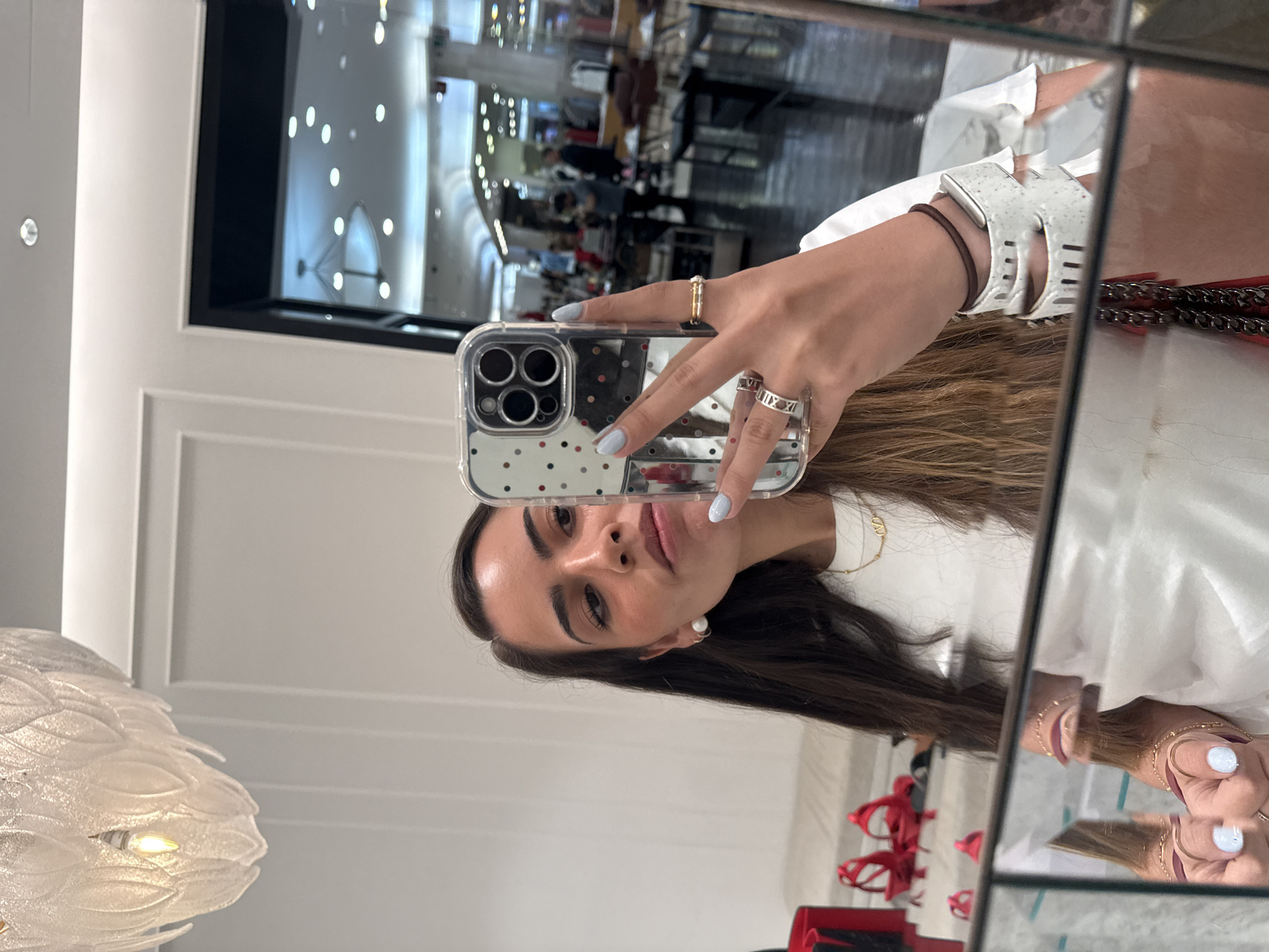 A woman is taking a selfie in a mirror inside a store. The store has a modern, high-end interior with white walls, a chandelier, and displays of merchandise. The reflection in the background of the mirror suggests the store is likely located in a shopping mall or commercial area. The woman is holding a smartphone with a patterned case, and her reflection is visible in the mirror. She is wearing a white shirt and has jewelry on her fingers. Without specific identifying details, pinpointing the exact location in Puebla, Mexico, is not possible.