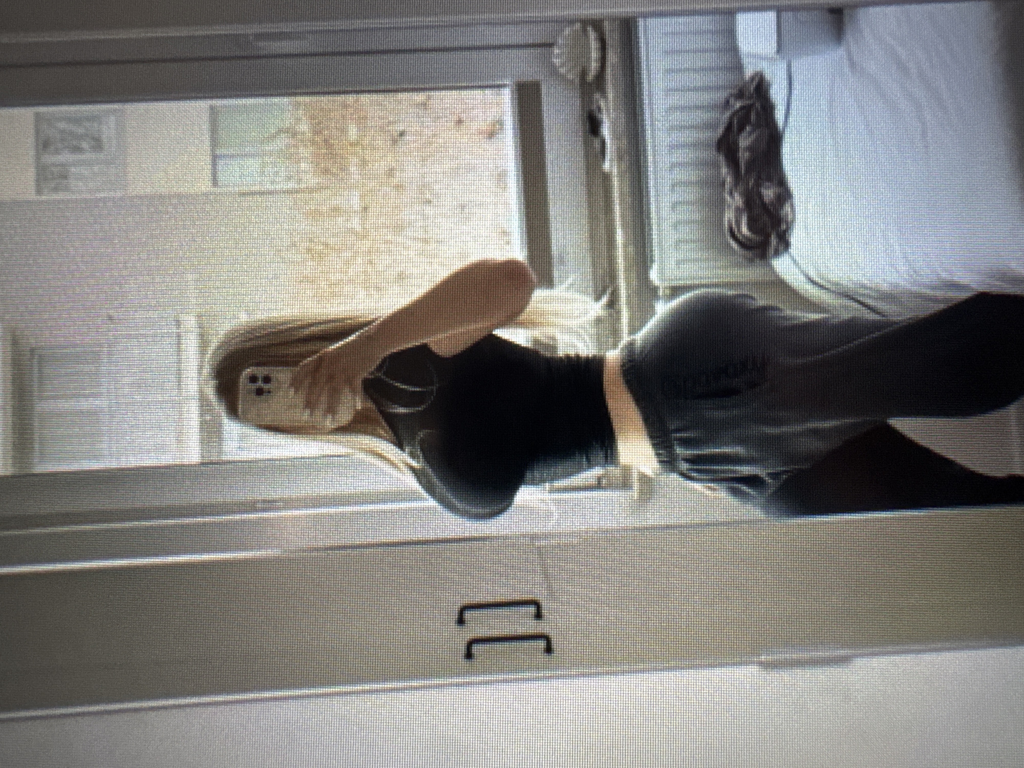 The image displays a person taking a mirror selfie indoors, captured from a sideways perspective (rotated 90 degrees clockwise).
**Subjects:** The main subject is a young person, likely female, with long blonde hair, standing in front of a mirror. Her face is obscured by a light-colored smartphone she is holding up to take the photo. She is wearing a fitted black long-sleeved top and dark-colored pants or leggings.
**Objects:** The smartphone has a light-colored case. The room appears to have a large window or glass door, through which an outdoor scene is visible, including bare trees or bushes, suggesting autumn or winter foliage, and possibly distant buildings. Inside the room, to the right of the person's reflection, a bed with white bedding is visible. A dark, crumpled piece of clothing or fabric rests on what appears to be a radiator or heating unit next to the bed. Below the person's reflection, two dark, U-shaped handles, possibly from a drawer or cabinet, are visible.
**Scene/Setting:** The setting is an indoor room, likely a bedroom or living area, with natural light entering from a large window or glass door. The person is reflected in a full-length mirror.
**Activity or event taking place:** The person is actively taking a mirror selfie using their smartphone.
**Notable details:** The time of day appears to be daytime, indicated by the natural light. The outdoor view of sparse trees suggests either late autumn or winter. The overall image quality is somewhat grainy or pixelated, potentially indicating it is a photo of a screen or taken with a lower resolution.
**Visible text:** There is no visible text in the image.