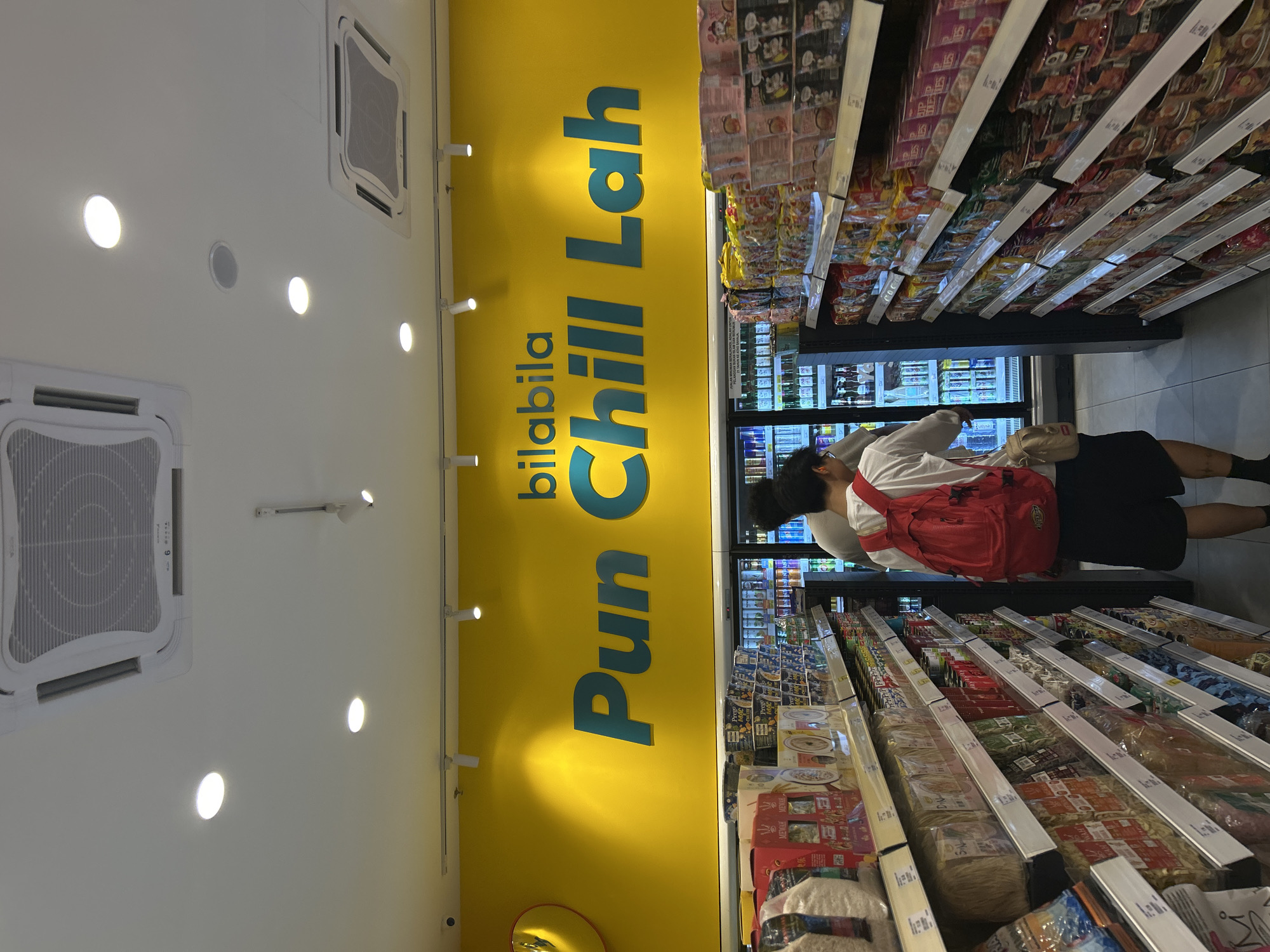 The image captures the interior of a retail space in Subang Jaya City Council, Malaysia. The store features shelves laden with packaged food items, running along the right side of the frame. A person wearing a white shirt, black shorts, and a red backpack is browsing the items. Behind the person is a refrigerated display case filled with various products. The left side of the frame shows a yellow wall with the text "bilabila Pun Chill Lah" in blue and white ceiling. The ceiling contains recessed lighting and square air conditioning vents.