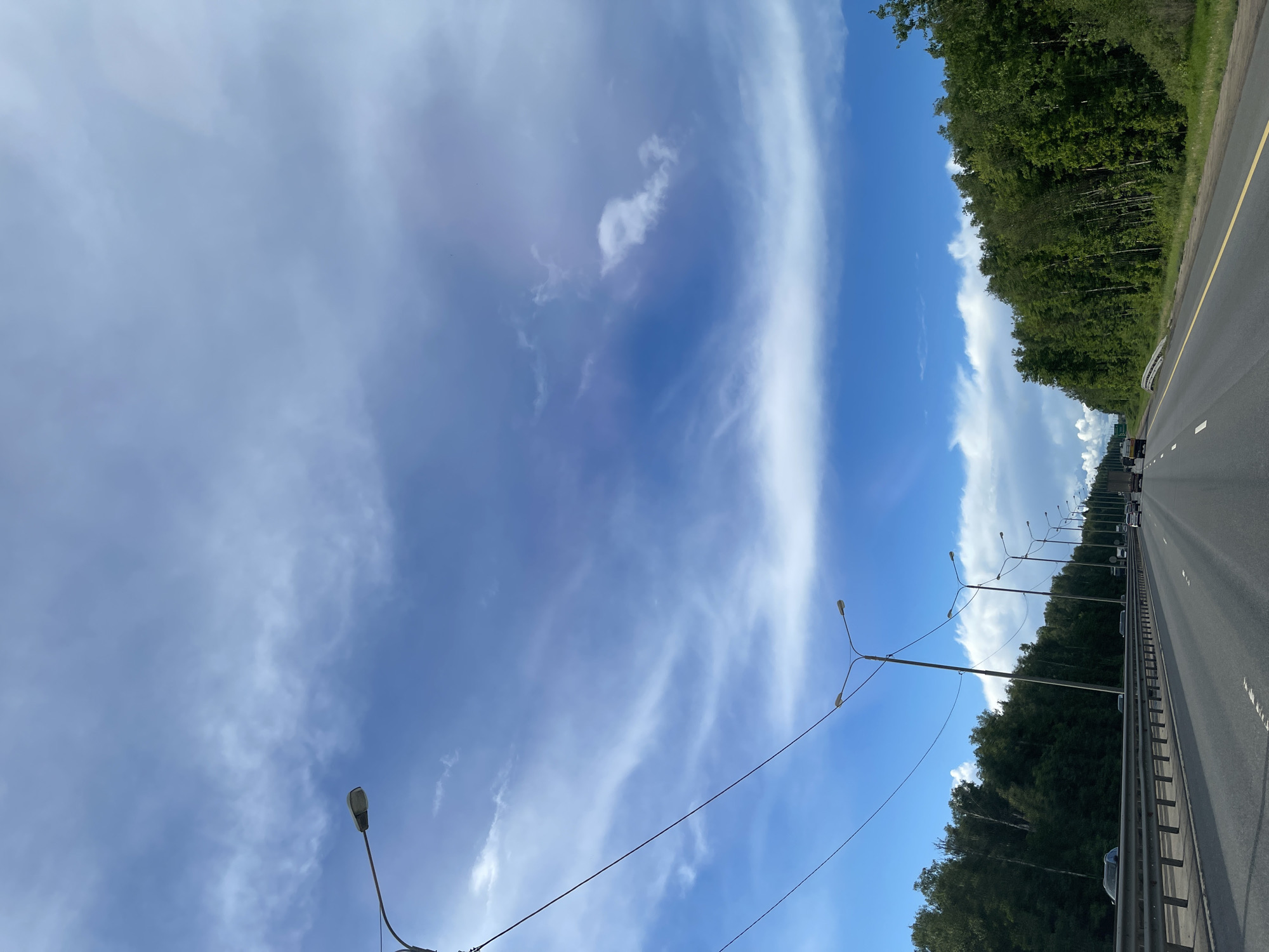 The image shows a view of a highway, likely captured from a moving vehicle. The scene includes a clear blue sky with wispy clouds, with a distinct separation of cloud formations. A row of street lights lines the side of the road, interspersed with trees on the embankment. The highway is divided by a metal barrier. Cars are visible on the roadway. Given the location is near Serp I Molot, Russia, it is possible that this scene depicts a highway in that region.