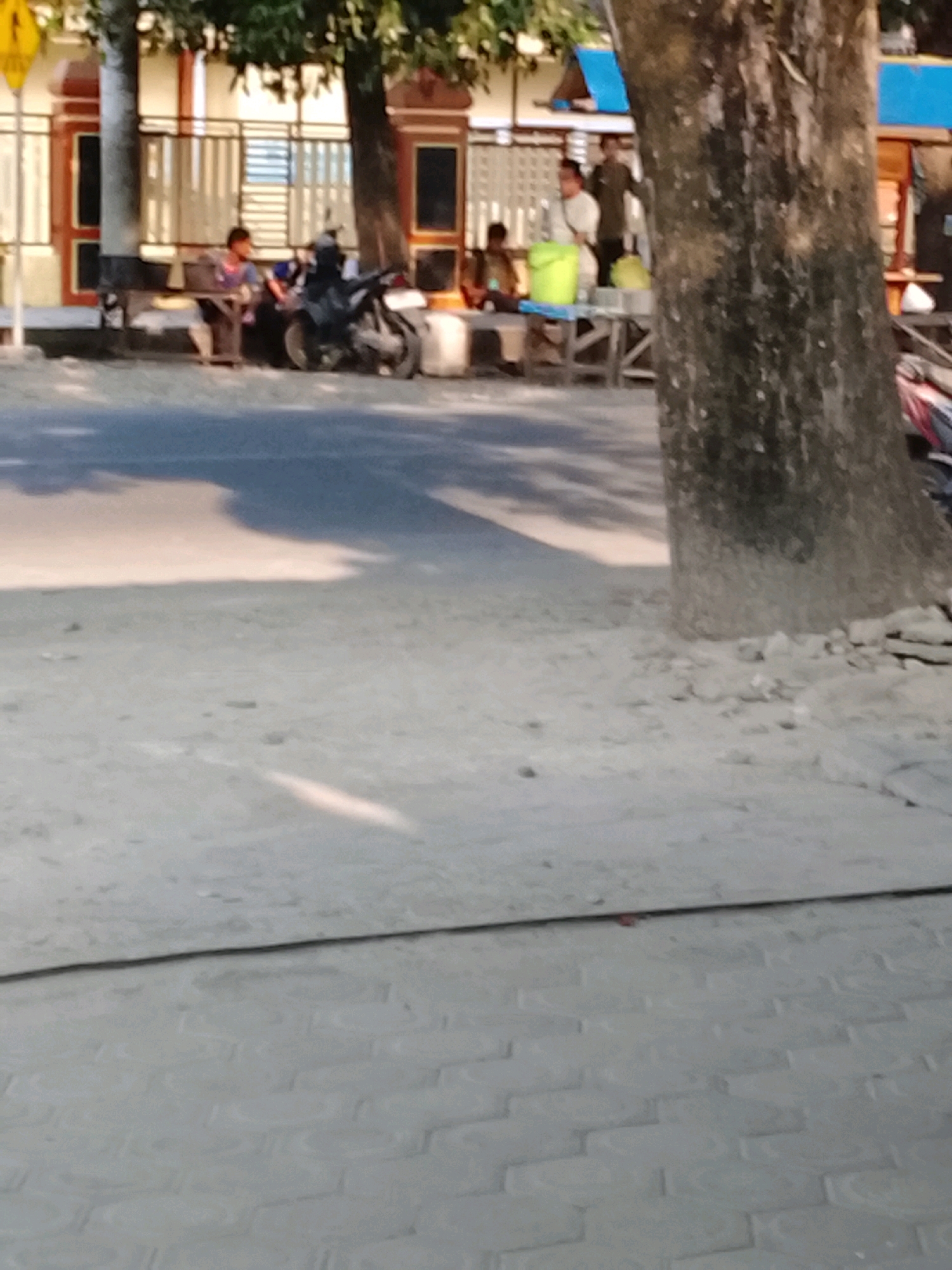 The image depicts an outdoor scene in Slawi, Indonesia, possibly near a roadside food stall or waiting area. In the foreground, a dusty, paved surface is visible, with a distinct line separating it from a brick-like pattern. A large tree trunk dominates the right side of the image, casting shadows onto the ground.
In the background, several individuals are present. One person, wearing a dark shirt, sits on a bench next to a parked motorcycle. Another individual, in a lighter colored shirt, is also seated. Further back, two people appear to be standing near a table with a yellow bucket on it. The area behind them suggests a building with decorative pillars and barred windows. The overall impression is of a casual, public gathering space, likely in a commercial or residential area of Slawi.