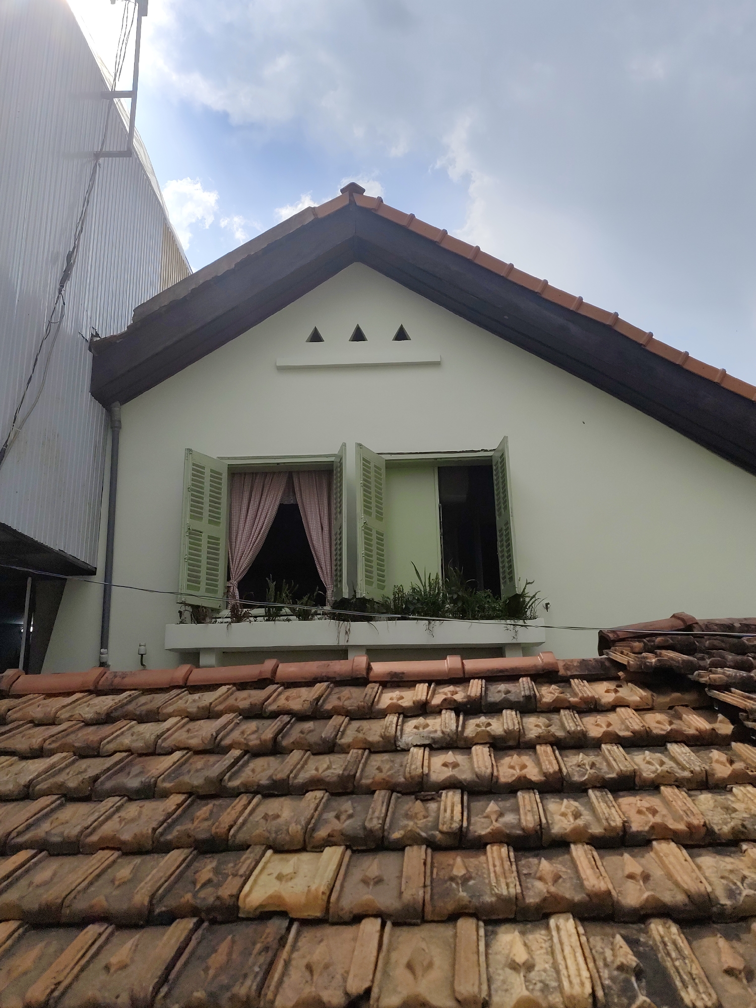 The image shows a building in Da Lat, Vietnam, as evidenced by the architectural style and roofing materials commonly found in the region. The foreground is dominated by aged, brown, interlocking clay roof tiles with a distinctive pattern. Behind this, a section of the building's facade is visible. It features a cream-colored wall with a gabled roofline trimmed in dark brown. Below the gable, there's a row of three triangular decorative vents. A window is centered on the wall, with open, light-green shutters and a small balcony or shelf below, containing plants. A red-and-white checkered curtain is visible through the window. To the left of the building, a gray corrugated metal structure extends upwards, reaching towards the sky, where clouds can be seen. A wire is strung across the scene, partially obscuring the facade.