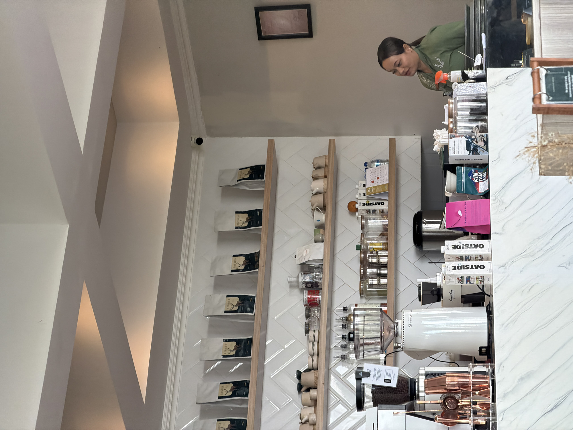 The image depicts the interior of a coffee shop, likely located in Baturiti, Indonesia, based on provided context not directly visible in the image. The scene is a counter area with shelves stocked with various coffee-related items. On the top shelf, several bags of coffee beans with a deer graphic are displayed. The middle shelf holds an assortment of small ceramic cups and jars, as well as bottles and a red mug. The lower shelf, behind the counter, displays more jars, coffee equipment including a grinder, a coffee maker, and various cartons, such as "OATSIDE." A female employee, wearing a green uniform with a deer emblem, is visible behind the counter, attending to something not fully in frame. The environment appears clean and well-organized, with a modern aesthetic characterized by light-colored walls and shelving. The lighting suggests an indoor setting, possibly during daytime. A security camera is mounted on the wall near the ceiling.
