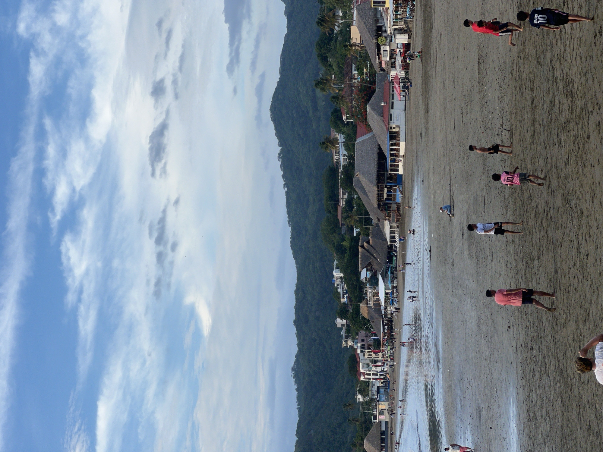 This is a vertical, eye-level, medium shot of a beach scene in San Juan del Sur, Nicaragua. The foreground is dominated by a sandy beach where several young males are playing what appears to be soccer. One individual in a pink shirt and dark shorts is facing away from the camera, while another in a white shirt and dark shorts is in the middle distance. In the far foreground, a person wearing a white shirt is visible from the chest up, with their arm raised in a gesture.
The middle ground of the image features a shallow body of water where several people are walking and playing. Beyond the water, a stretch of beachfront buildings, many with thatched roofs and open-air seating, are visible. These structures appear to be businesses such as restaurants or shops. In the background, a lush, green, tree-covered hill rises steeply, with more buildings sparsely scattered on its slopes. The sky above is a mixture of blue and white clouds. The overall impression is of a lively beach town during daylight hours, with recreational activities taking place both on the sand and in the shallow water.