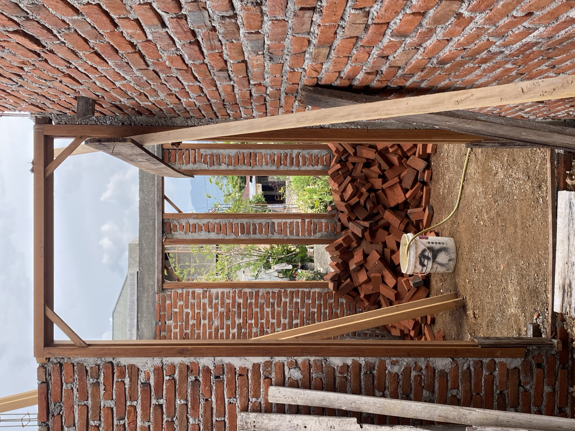 In an outdoor construction site, a partially built brick structure with a wooden frame stands under a cloudy sky. The foreground on the left shows a stack of wooden planks. To the right, a brick wall extends into the distance. The central area of the image reveals a doorway-like opening framed by wood, with a large pile of red bricks scattered on the dirt floor within. A white bucket with a yellow hose coiled around it is situated among the bricks. In the background, glimpses of vegetation and other buildings can be seen, with distant hills under a hazy sky.
