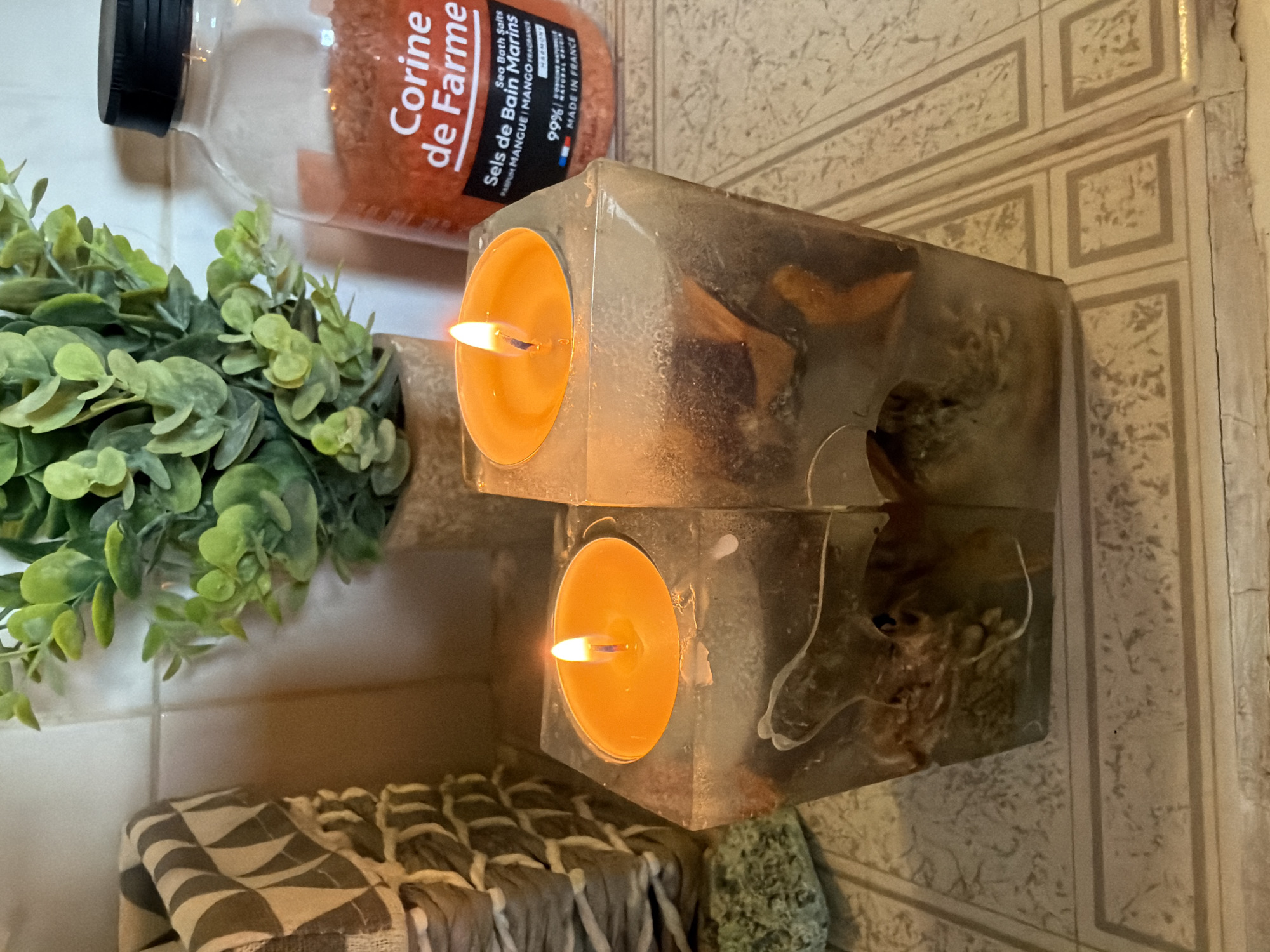 This media file shows a close-up, top-down view of a bathroom or spa-like setting. The primary subjects are two clear, rectangular candles with orange tea lights burning inside, placed on a tiled surface. To the left, a clear bottle filled with orange bath salts is visible, along with a faux green leafy plant. In the foreground, a folded towel with a geometric pattern and a small green textured object are present. The lighting suggests a dim, ambient environment, likely indoors, possibly in the evening or a room with low natural light. The visible text on the bath salt bottle reads: "Corine de Farme," "Sea Bath Salts," "Sels de Bain Marins," "PARUM MANGUE/MANGO FRAGRANCE," "99% NATURAL ORIGIN," "HARMONY," and "MADE IN FRANCE." The overall impression is one of relaxation and self-care.