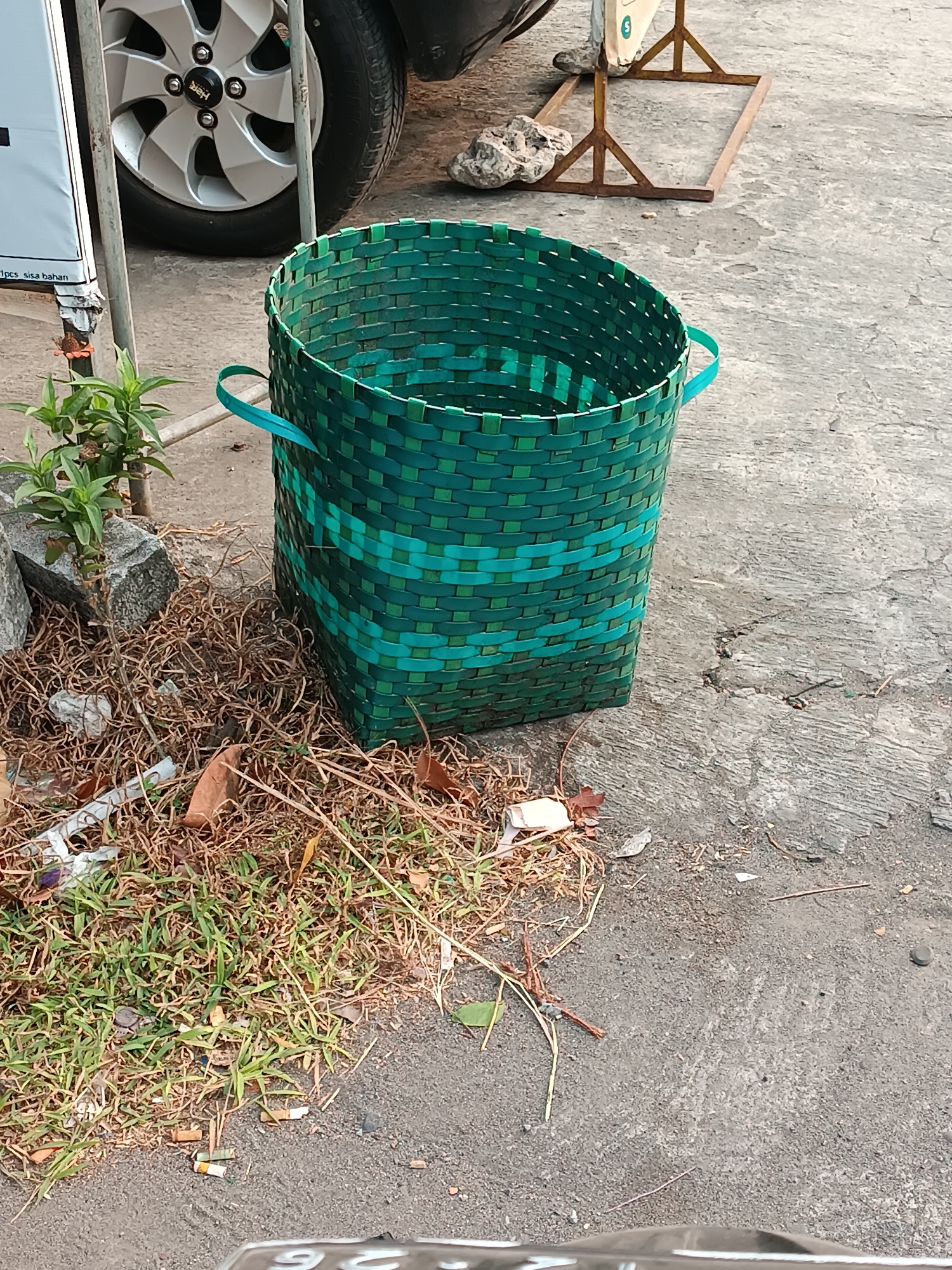 The image depicts an outdoor scene in Pleret, Indonesia. In the foreground, a green woven basket sits on a concrete surface littered with dry grass, leaves, and discarded cigarette butts. A small plant with green leaves grows from behind a grey rock next to the basket. To the left, a portion of a white sign with black text is visible, along with a metallic pole. Behind the basket, the silver wheel of a vehicle is partially visible. In the background, a rusty metal structure stands on the concrete. The overall environment appears to be an urban or semi-urban area, with a focus on discarded items and a woven basket as the central subject.