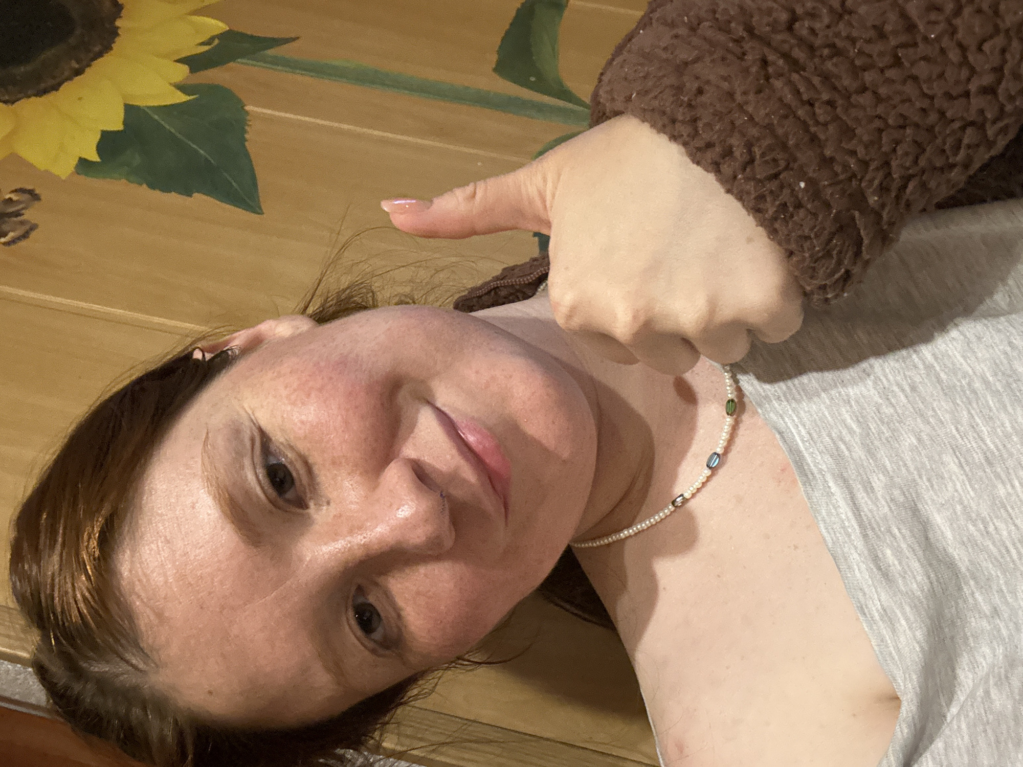 A woman with fair skin, reddish-brown hair, and freckles is angled towards the viewer, giving a thumbs-up gesture. She wears a light gray t-shirt and a pearl necklace with interspersed colored beads. Her right arm is covered in a fuzzy brown material. The background is a wooden surface with a painted sunflower design. There is no information in the image to indicate a location in the city of Nizhny Novgorod, Russia.