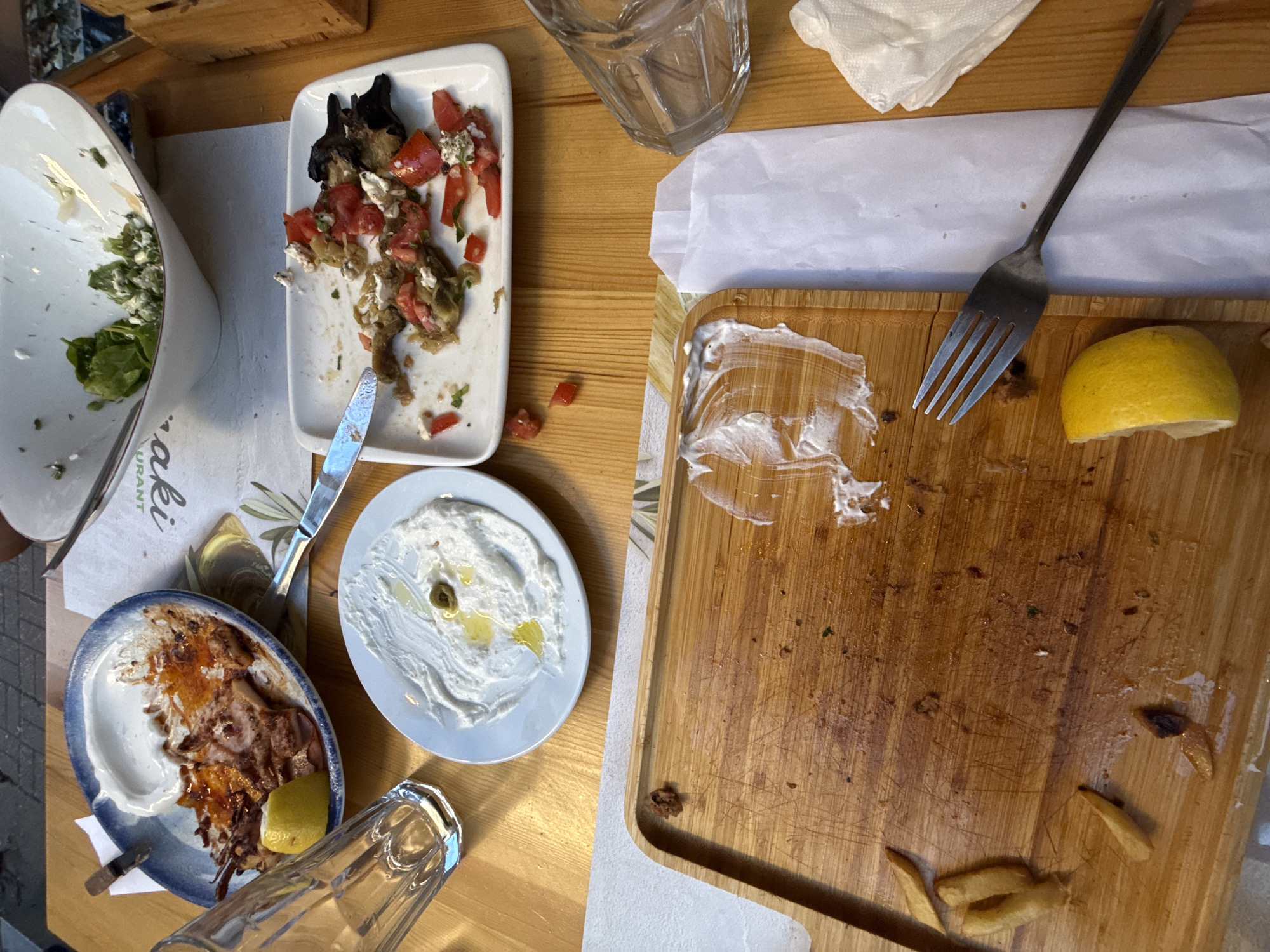 The image depicts a wooden table at an outdoor restaurant in Plovdiv, Bulgaria, after a meal. A partially eaten dish of grilled octopus with lemon and a smear of yogurt or dip is on a blue and white plate. Next to it is a white rectangular plate with remnants of roasted vegetables, tomatoes, and feta cheese, with a knife resting on it. Another small white plate contains a dollop of white dip with a single olive and a drizzle of olive oil. A wooden cutting board in the foreground is scattered with a few french fries, a lemon wedge, and streaks of white sauce. Two empty drinking glasses are visible, one to the left and another to the right. A white napkin and a white paper wrapper are also on the table. In the background, a large white bowl contains a small amount of salad greens and crumbled cheese. The overall scene suggests the conclusion of a meal.