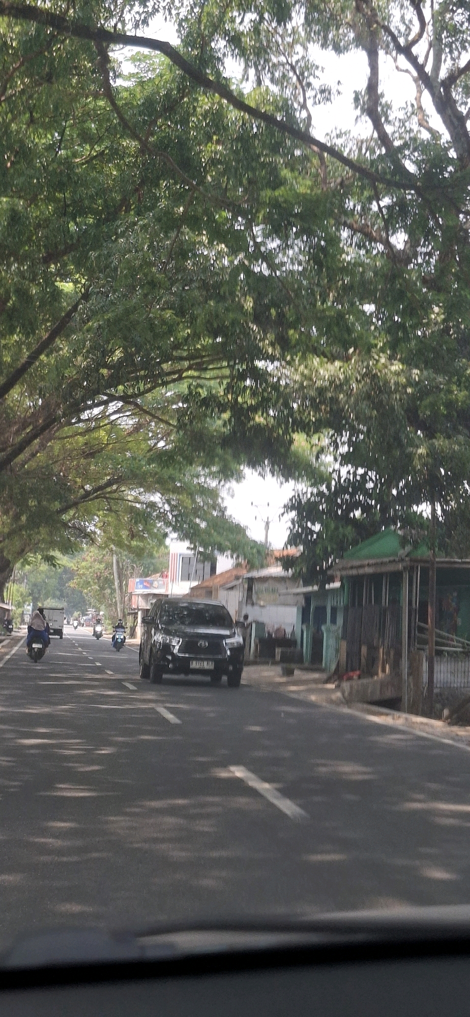 The image depicts a street scene in Susukan, Indonesia, characterized by a road lined with mature trees creating a canopy overhead. On the paved road, a black SUV is traveling in the right lane, its license plate partially visible. To the left, several motorcycles are also in motion. One motorcycle in the left lane has a rider wearing a blue headscarf. Further down the road, other vehicles, including a truck, are visible. The roadside features buildings with a mix of architectural styles, some appearing to be shops or residences, with one structure having a green roof. The overall environment suggests a sunny day with dappled sunlight filtering through the trees onto the road and surrounding area.