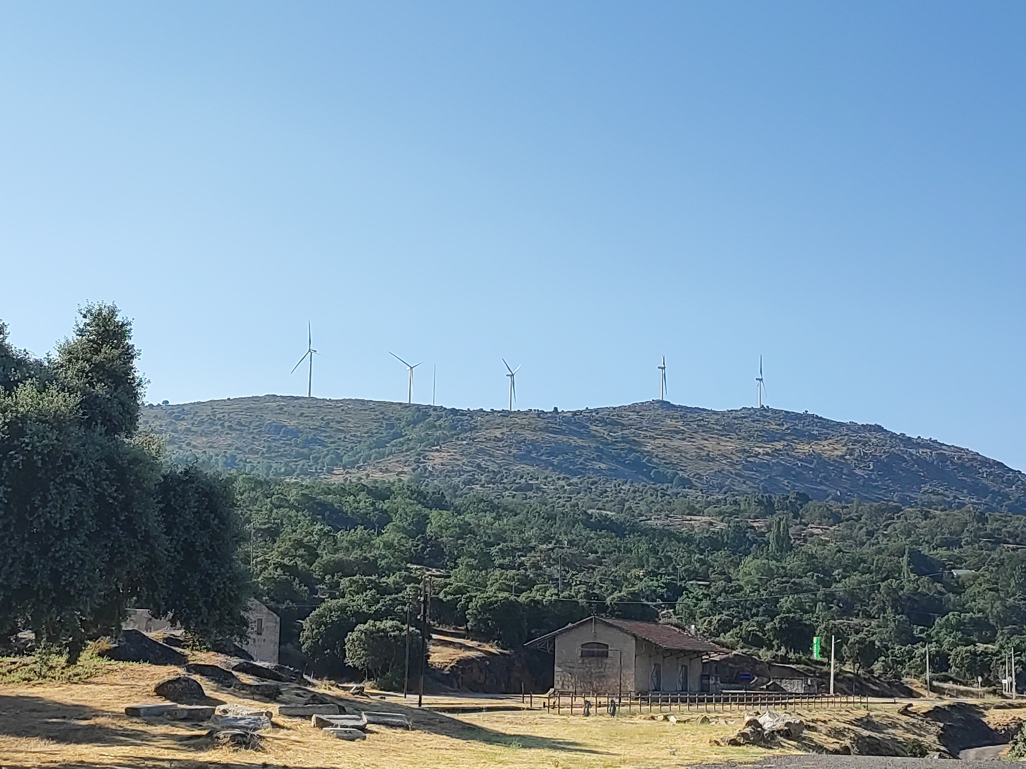 The image captures a pastoral landscape on a clear, sunny day, with a distinct presence of wind turbines atop a rolling, tree-covered hill. In the foreground, a rustic, stone building with a tiled roof stands prominently, suggesting a historical or rural structure, possibly an old railway station or an industrial building. The surrounding area is characterized by dry, yellow grass and scattered rocks, with a dense expanse of green trees covering the lower slopes of the hill. Several wind turbines are visible on the crest of the hill, silhouetted against the bright blue sky. The scene evokes a sense of serene, undeveloped nature, with a touch of human technological intervention. The location is identified as Ledrada, Spain, hinting at the geographical context of this tranquil scene.