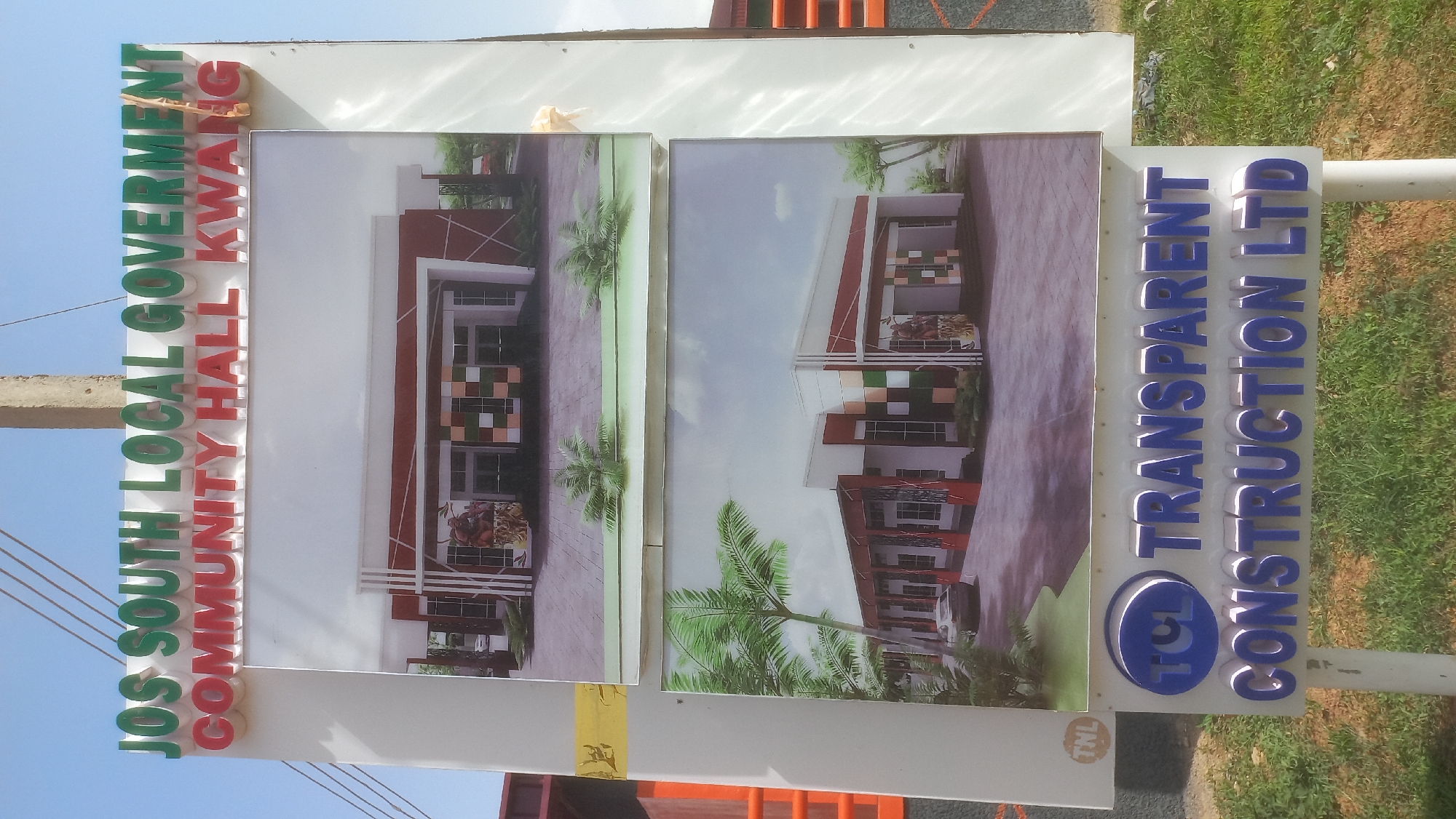 This is a vertical, full-shot photograph of a sign for the Jos South Local Government Community Hall Kwang, located in Jos, Nigeria. The sign features two architectural renderings of a modern community hall, adorned with palm trees and a cloudy sky. The text "JOS SOUTH LOCAL GOVERNMENT COMMUNITY HALL KWANG" is displayed vertically in large, colorful letters on the left side of the sign, with "JOS SOUTH LOCAL GOVERNMENT" in green and "COMMUNITY HALL KWANG" in red. On the right side of the sign, the text "TRANSPARENT CONSTRUCTION LTD" is written in blue, dimensional lettering, accompanied by the TCL logo. The sign appears to be illuminated, suggesting it may be visible at night. The overall scene depicts a sunny day, with the sign positioned outdoors, next to a grassy area and a white pole. The image is slightly angled, giving a sense of depth and perspective to the scene. There are no people visible in the photograph.