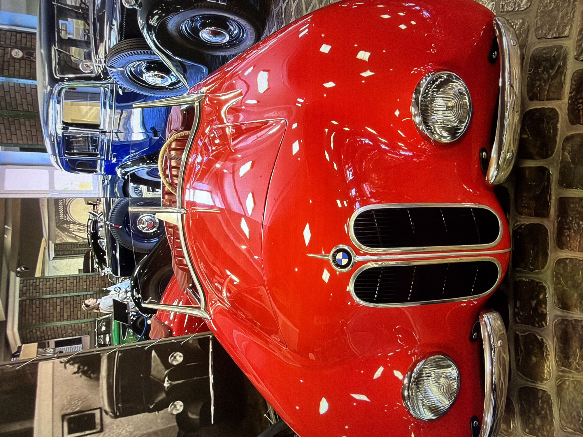 The image shows the interior of a car museum, possibly the Russian State Museum of Automotive History, located in Kotelniki, Russia. In the foreground, a bright red vintage convertible sports car is prominently displayed. Its polished hood reflects the surrounding environment. The car features a chrome grille with the BMW logo at the center, two round headlights, and chrome bumpers. The interior of the car is visible, showing red leather seats and a wooden steering wheel.
Behind the red car, other vintage vehicles are parked. To the left, a black vintage car is partially visible, with its headlights and grille showing. Further back, a dark blue vintage sedan is parked, with its spare tire mounted on the side. A woman in a grey top is standing to the left of the blue car, looking towards the right. The background shows brick walls and architectural elements, suggesting the building's interior. The floor is covered with what appears to be cobblestones. The overall impression is of a well-lit exhibition space dedicated to classic automobiles.