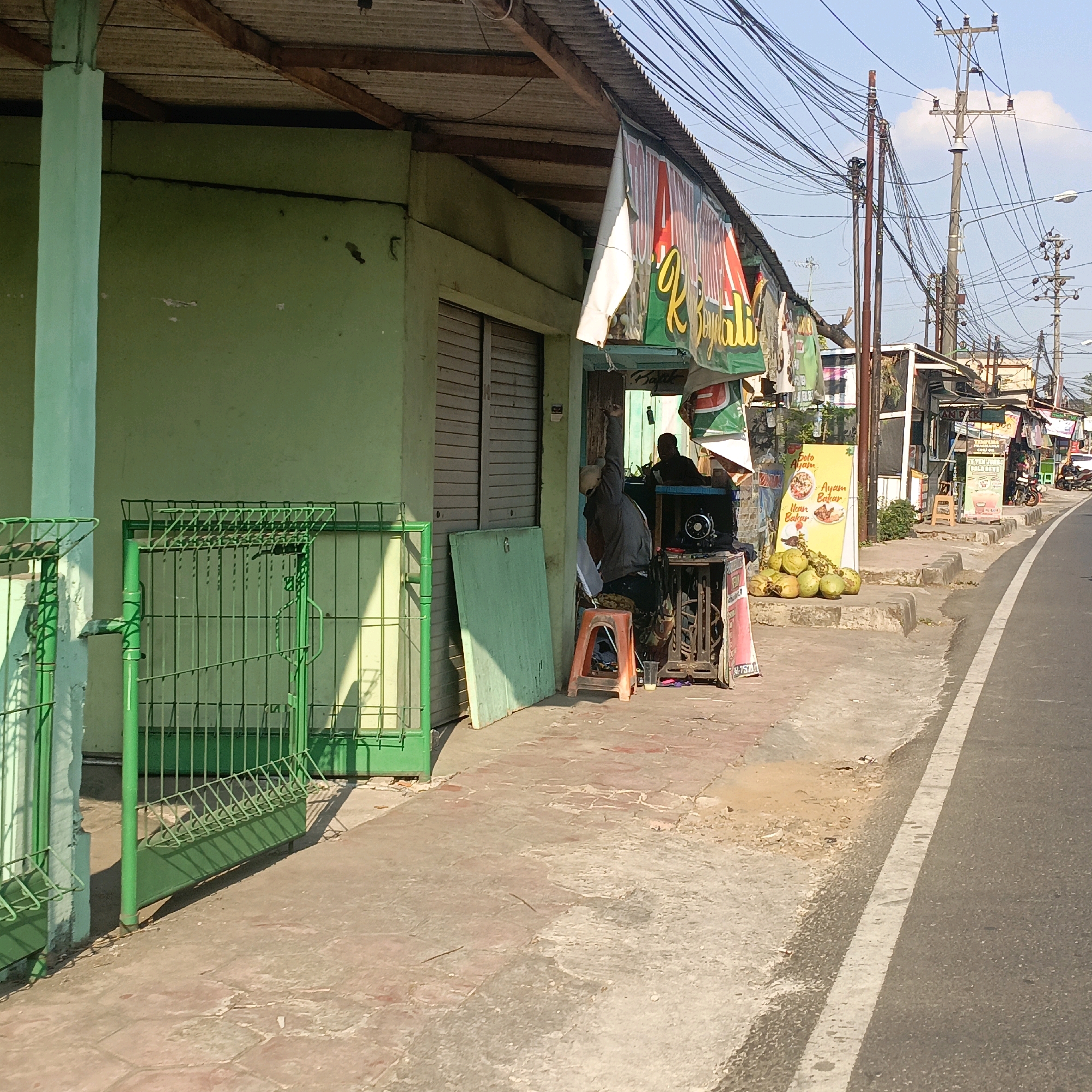 This is a street-level, eye-level, medium shot of a small roadside stall in Salatiga, Indonesia, on a sunny day. The stall is situated on the sidewalk next to a road. A green metal fence is in the foreground on the left. The stall itself appears to be part of a larger structure with a green exterior wall and a corrugated metal roof supported by wooden beams. A banner with red and green lettering, partially obscuring the building's facade, hangs over the stall area.
Two individuals are visible at the stall. One person, wearing a grey hoodie and a cap, is seated on an orange plastic stool and has their right arm raised. To their right, another person, wearing dark clothing, is seated behind a sewing machine that is mounted on a wooden table. A black object, possibly a speaker or another piece of equipment, is on top of the table. In front of the stall, a pile of green coconuts is stacked on a concrete base, next to a yellow sign with Indonesian text and images of food items like "Soto Ayam" and "Ayam Bakar." Further down the sidewalk, there are other small stalls and structures, with utility poles and a tangle of electrical wires overhead. The road in the background has a white lane marker and appears to be a moderately trafficked area, with a motorcycle visible in the distance. The sidewalk is paved with concrete tiles. The scene depicts a typical, functional commercial area in a city.