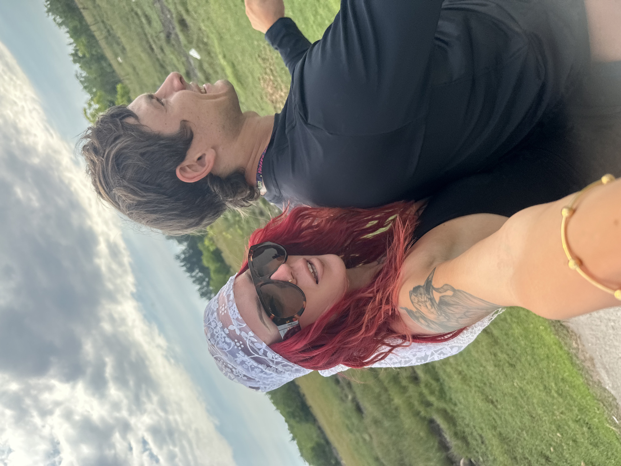 A photo captures two people outdoors in a grassy area. One person is lying down with the other person on top of them. The person lying down has short brown hair and wears a dark long-sleeved shirt and a necklace. The person on top has red hair, sunglasses, and a white lacy head covering, and has a bird tattoo on their upper arm, and also wears a gold bracelet. The location is a grassy field, the city and region are unknown.
